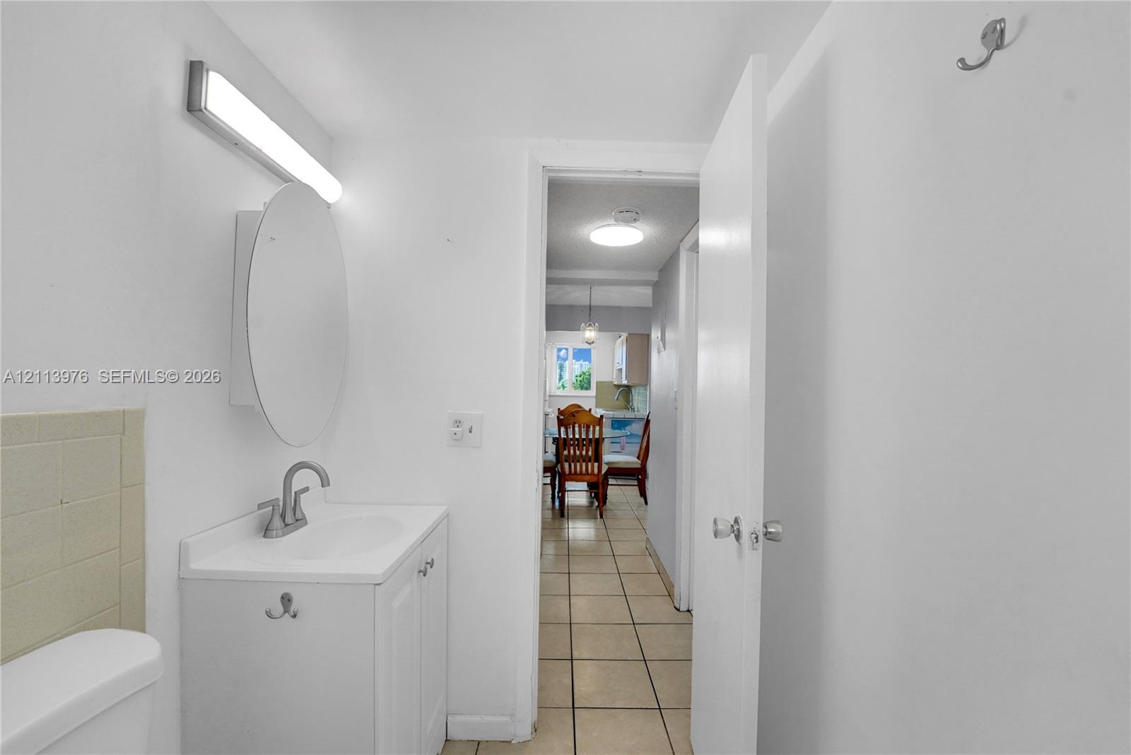 White vanity with integrated sink, circular wall-mounted mirror, linear light fixture, ceramic tile flooring, and wall-mounted hardware