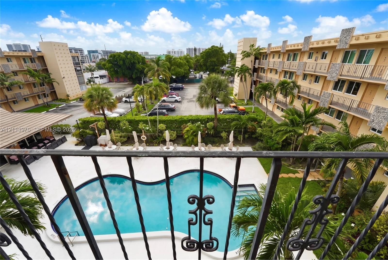 Curved swimming pool, concrete pool deck, wrought iron balcony railing, mature palm trees, and multi-story building exterior