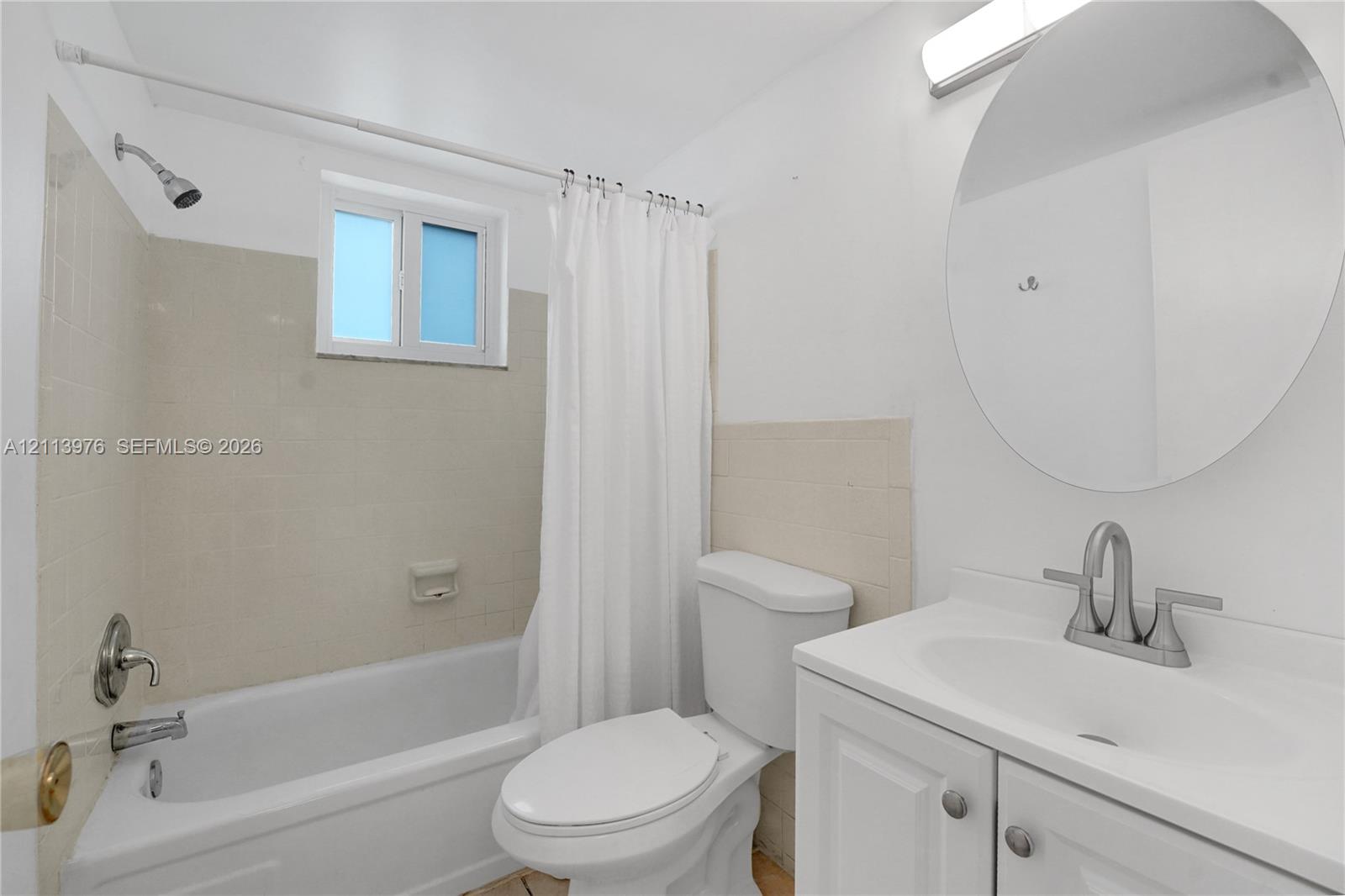 Bathroom featuring a white vanity cabinet with a integrated sink, brushed-nickel faucet, circular wall-mounted mirror, and a tub-shower combination with tile surround