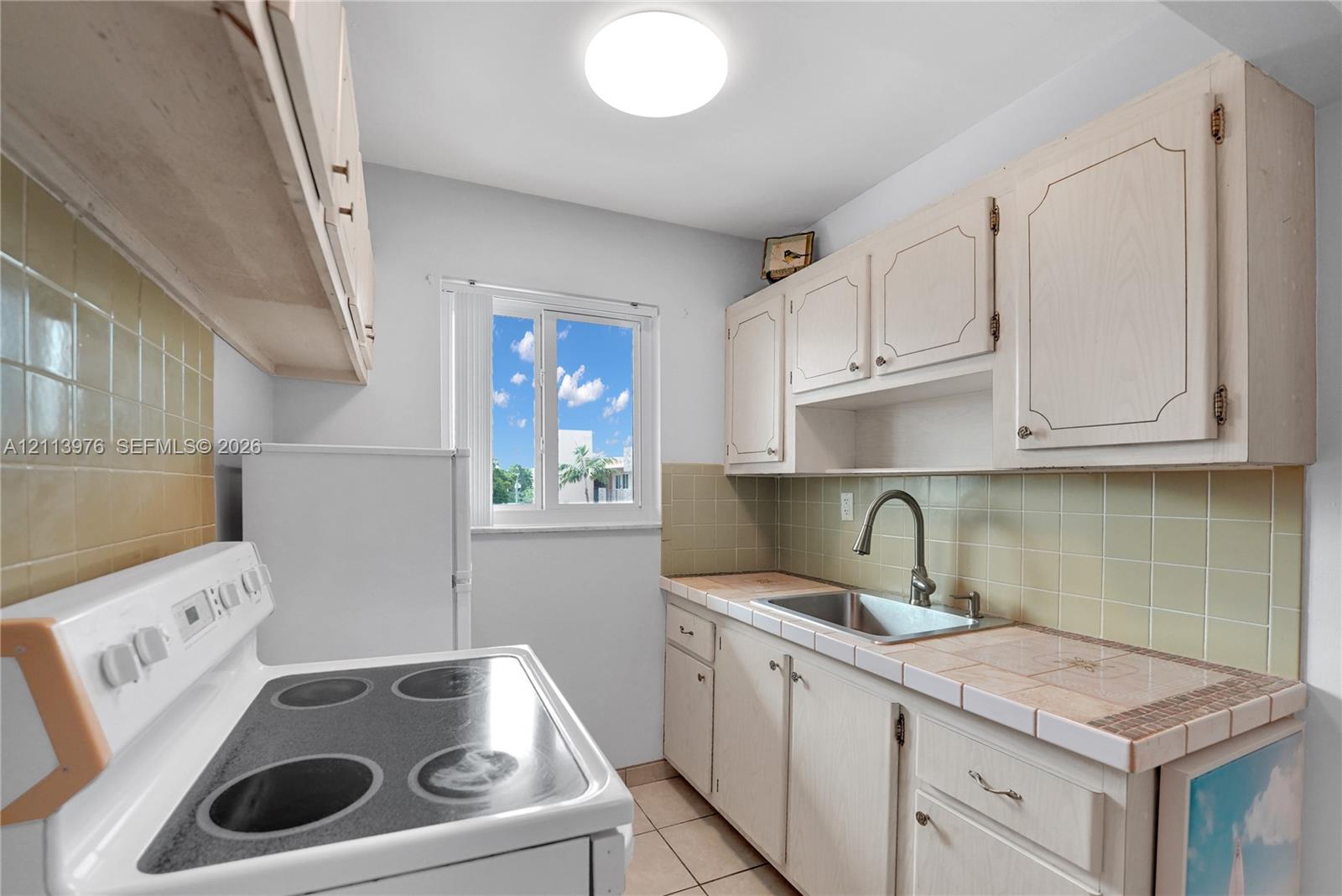 Kitchen featuring light-toned cabinetry, tile countertops, tile backsplash, stainless steel sink with high-arc faucet, and a flush-mount ceiling light