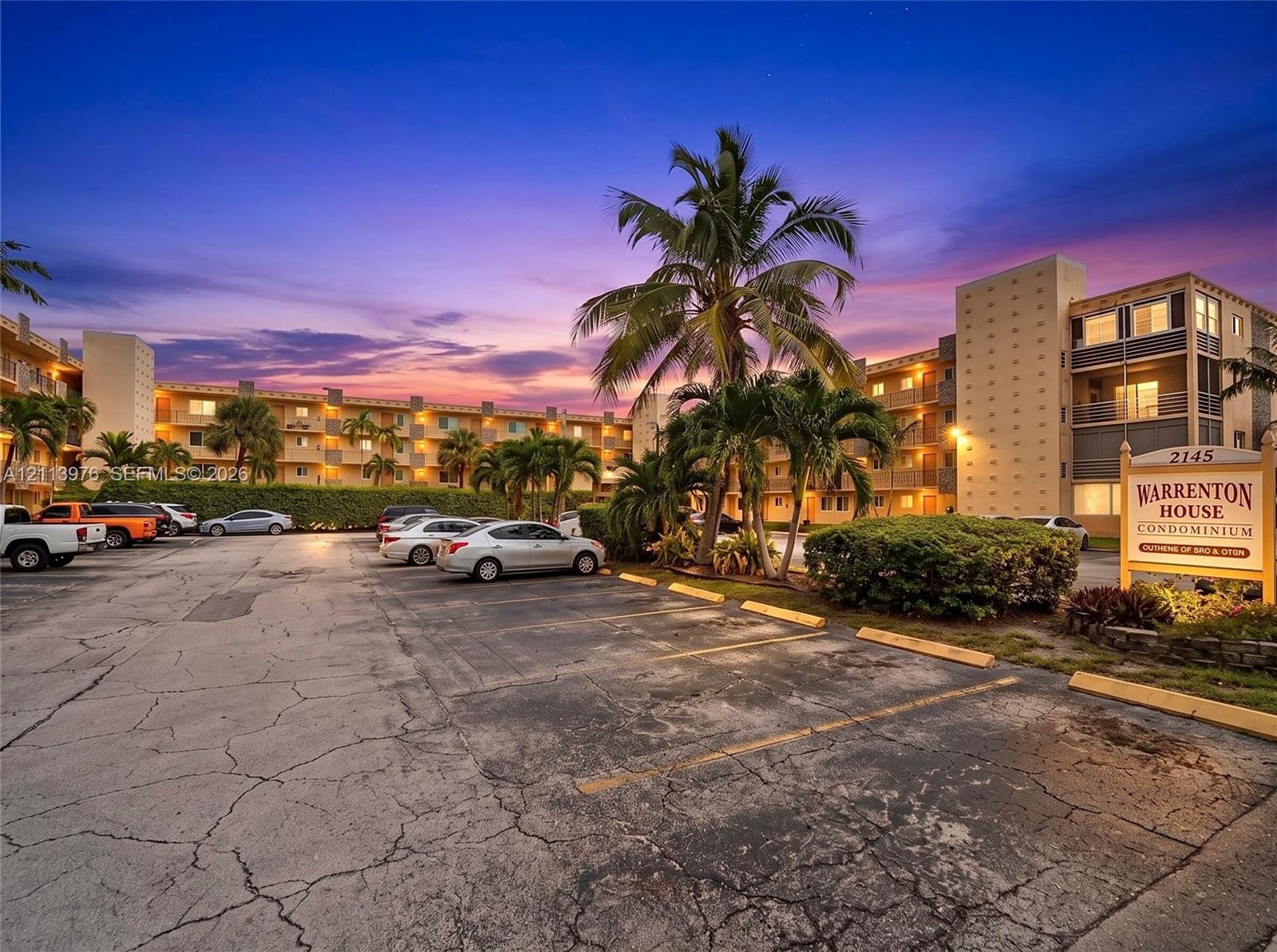 Multi-story condominium exterior featuring an on-site parking area, structured balconies, and mature palm trees