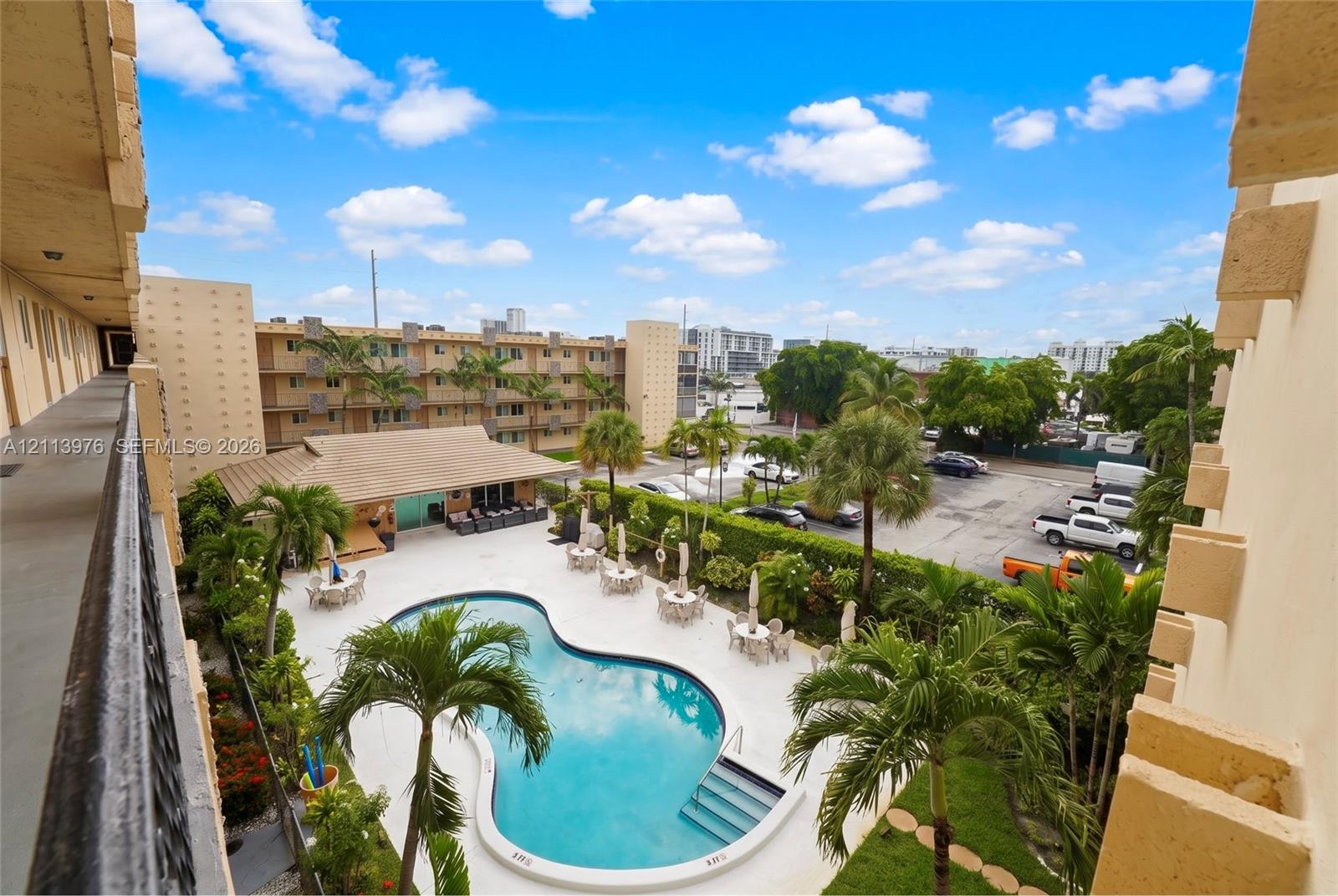 Community pool area featuring a free-form swimming pool, paved sundeck, lush landscaping with mature palm trees, and an attached covered cabana