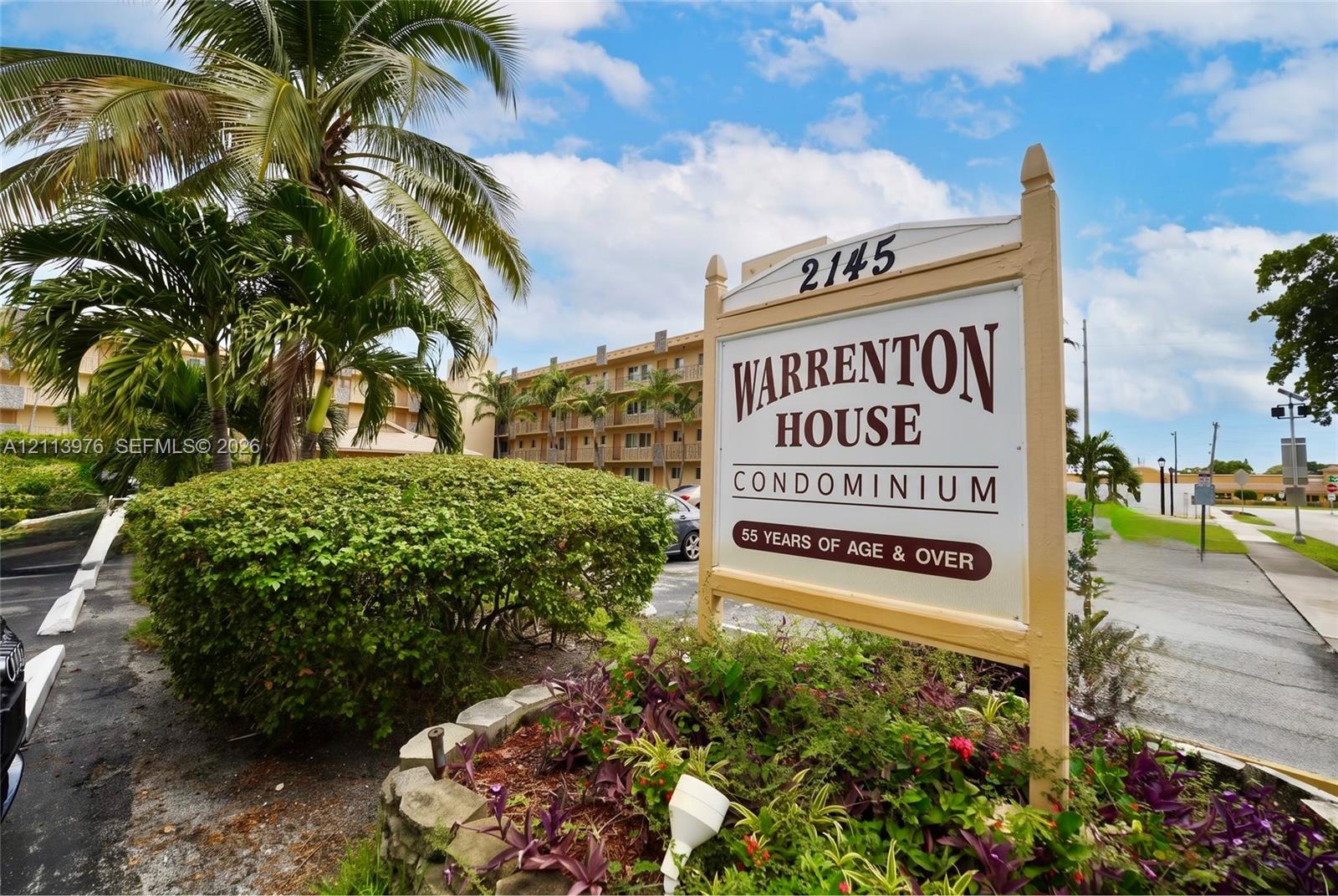 Multi-story condominium exterior featuring exterior balconies, mature palm trees, and landscaped stone-bordered garden beds