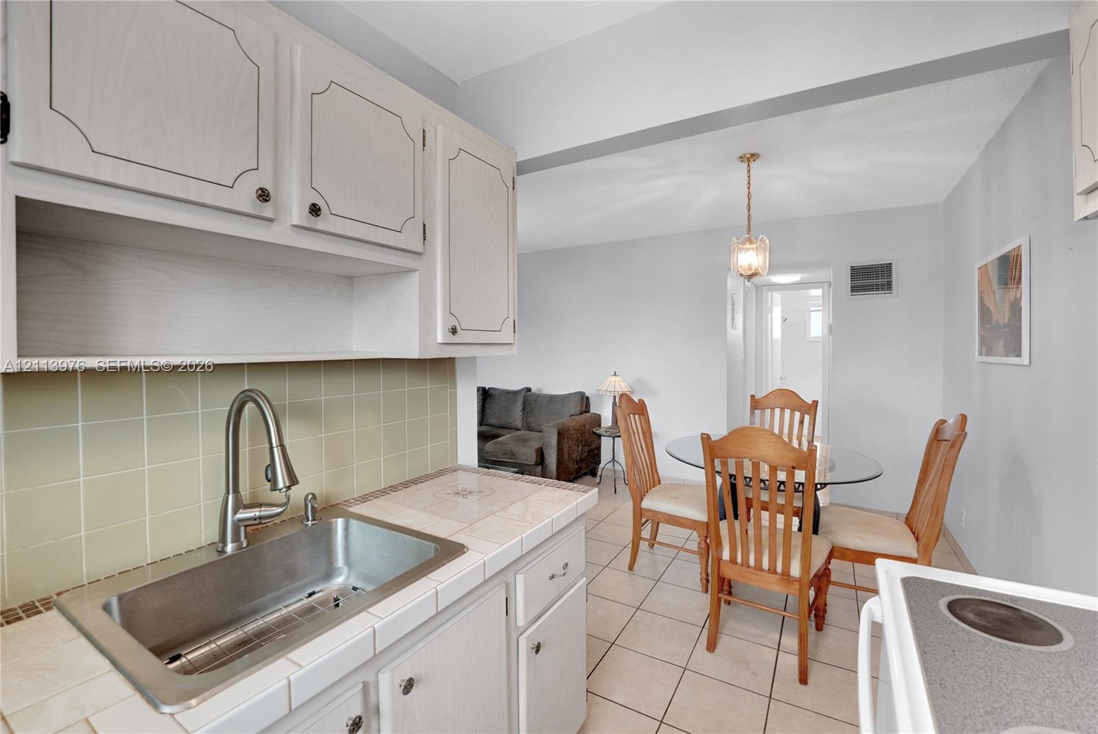 Kitchen featuring light-toned cabinetry, tile countertops, stainless steel sink with high-arc faucet, and ceramic tile flooring