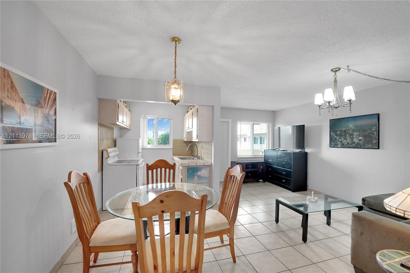 Kitchen area featuring tile countertops, white cabinetry, and a single-basin stainless steel sink, complemented by neutral tile flooring throughout and dual multi-arm metal chandeliers