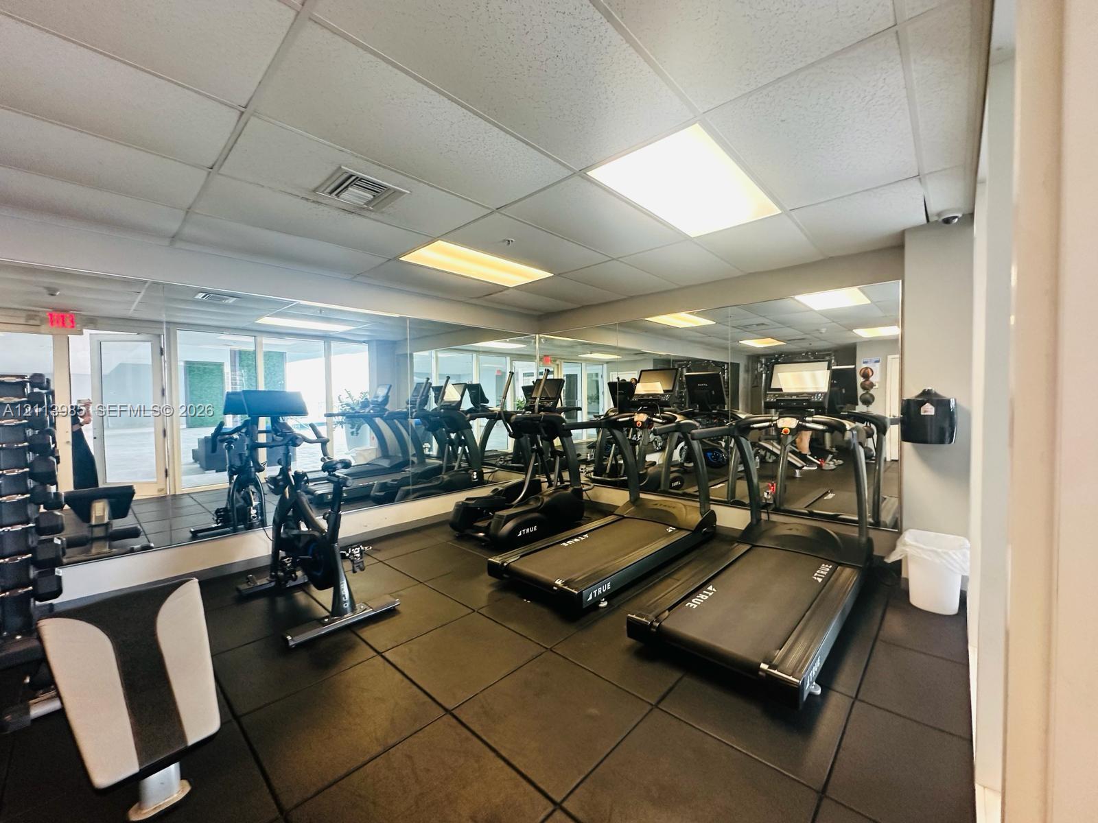 Fitness area featuring rubberized flooring, expansive wall-to-wall mirrors, recessed drop-ceiling lighting, and glass paneled doors