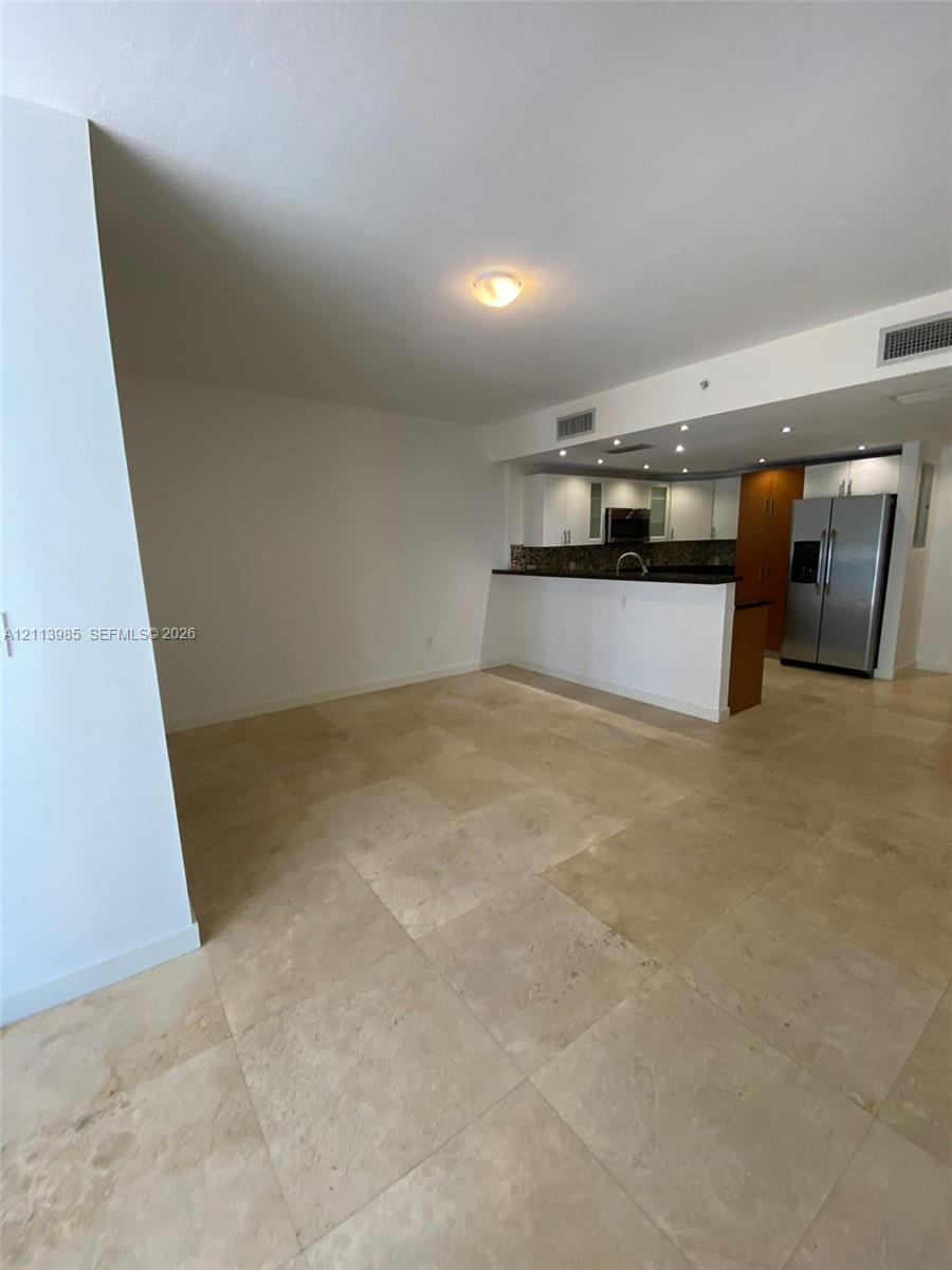 Large-format stone tile flooring, open kitchen peninsula with dark stone countertops, stainless steel refrigerator, and recessed ceiling lighting