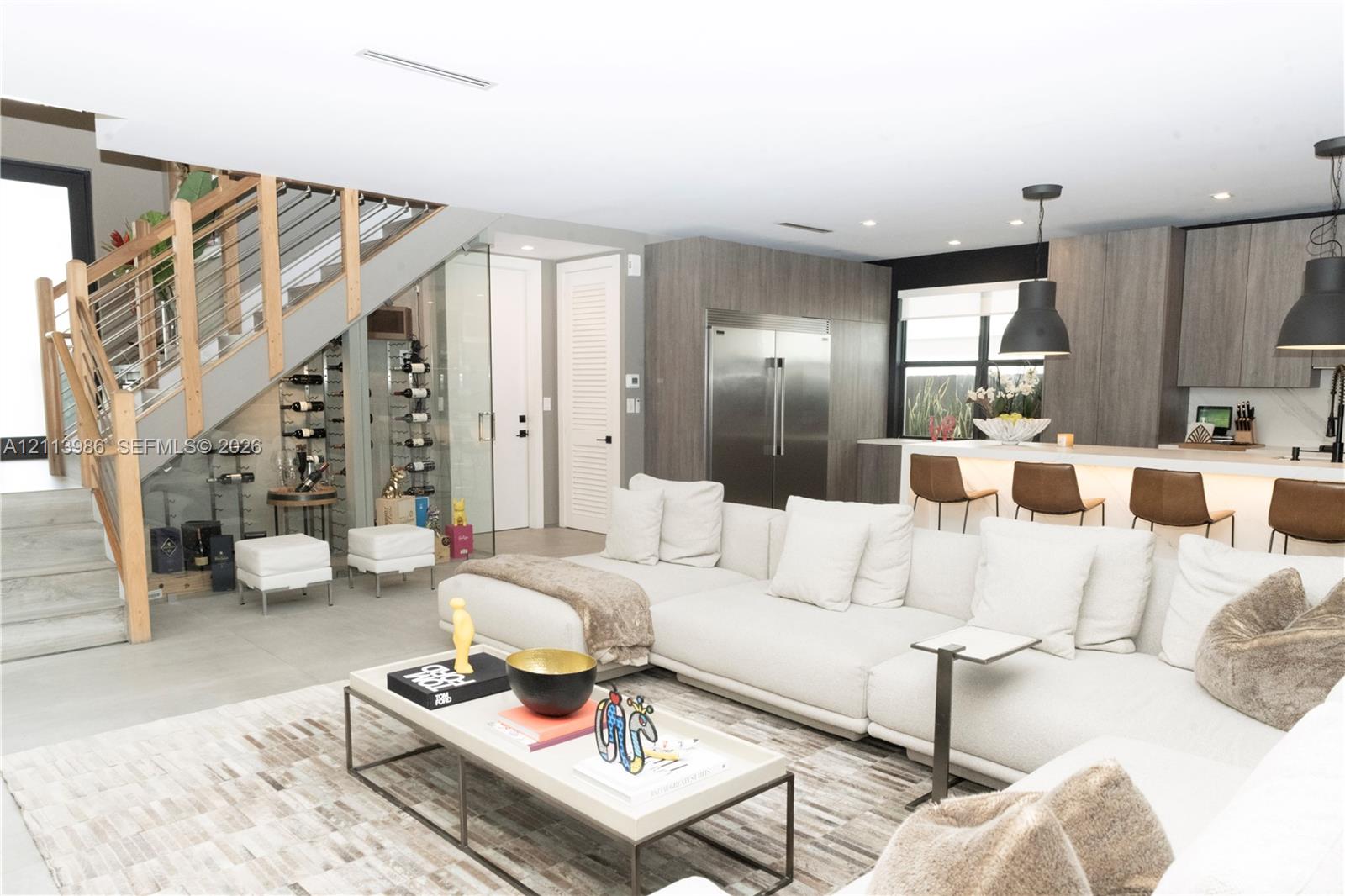 Open-concept kitchen featuring flat-panel cabinetry and stainless steel appliances, glass-enclosed wine storage, and a cable-railing staircase with wood handrails Open-concept kitchen featuring flat-panel cabinetry and stainless steel appliances, glass-enclosed wine storage, and a cable-railing staircase with wood handrails