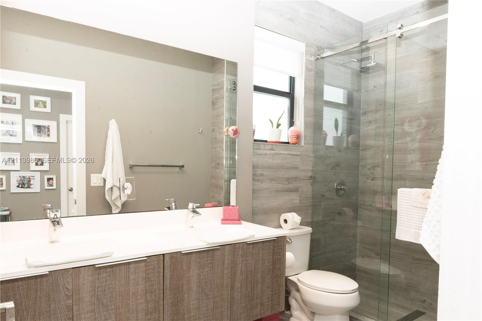 Dual-sink vanity with a white countertop and wood-finish cabinetry, frameless glass shower enclosure, and grey wood-finish tile shower wall