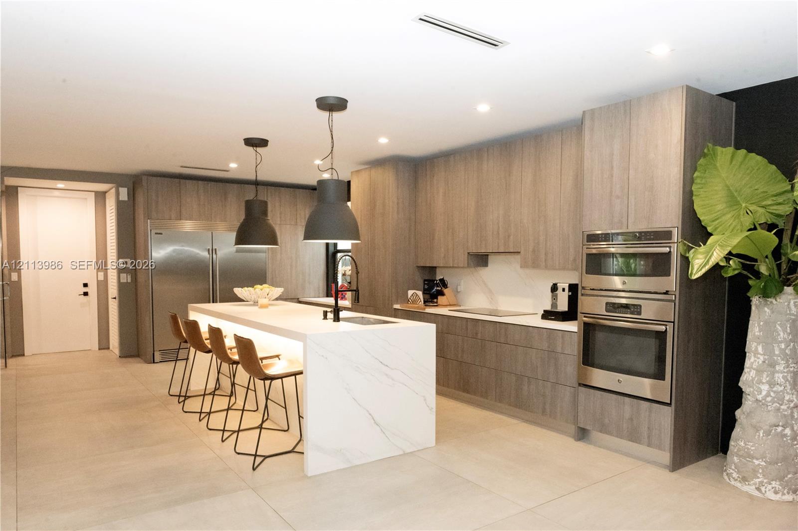 Kitchen featuring wood-finish flat-panel cabinetry, a waterfall island with stone countertop, stainless steel built-in double wall ovens, stainless steel refrigerator, and recessed lighting