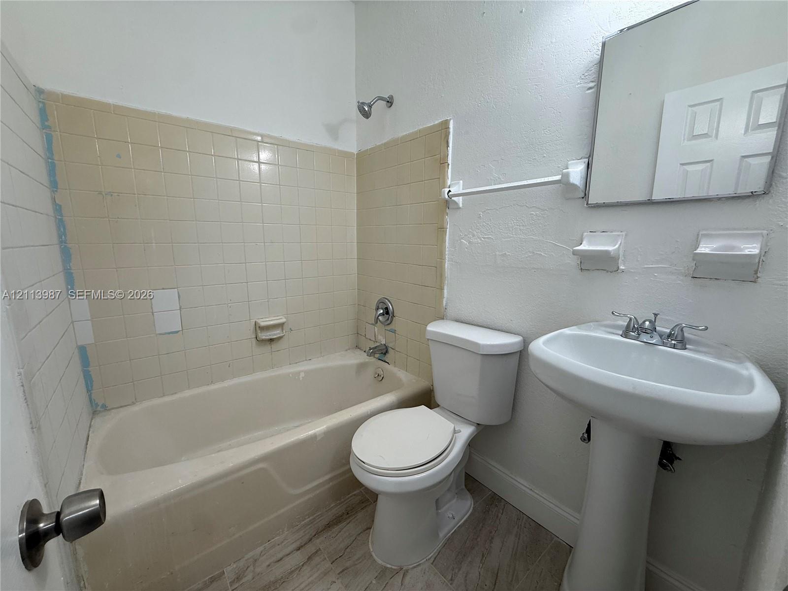 Bathroom featuring a pedestal sink, a bathtub with a tiled surround, a wall-mounted mirror, and wood-finish flooring Bathroom featuring a pedestal sink, a bathtub with a tiled surround, a wall-mounted mirror, and wood-finish flooring