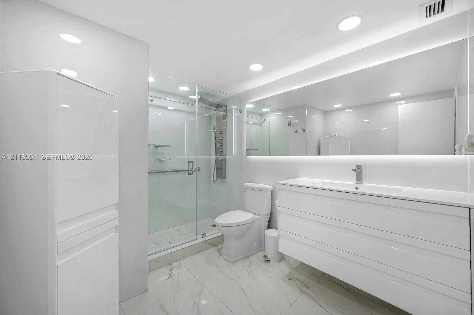 Modern bathroom featuring large-format tile flooring, a glass-enclosed shower with a multi-head body spray system, a floating vanity with a seamless integrated sink, and an expansive wall-to-wall mirror with integrated LED lighting