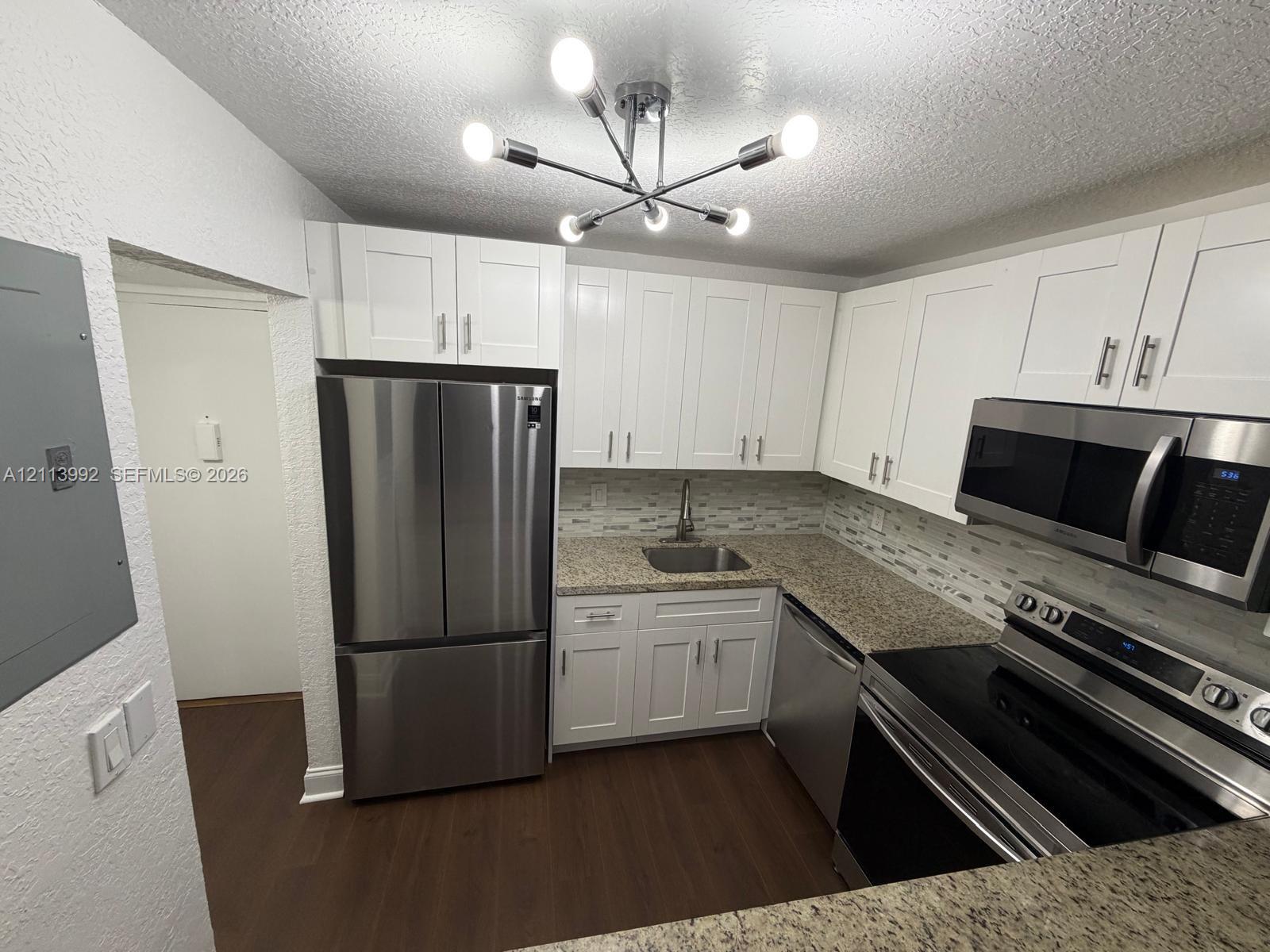 Kitchen featuring white shaker-style cabinetry, stone-finish countertops, stainless steel appliances, multi-toned tile backsplash, and a contemporary multi-arm light fixture