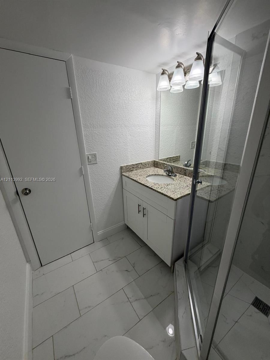 Bathroom featuring white cabinetry, stone-finish countertop with integrated oval sink, glass-enclosed shower, polished light-colored tile flooring, and multi-bulb vanity lighting