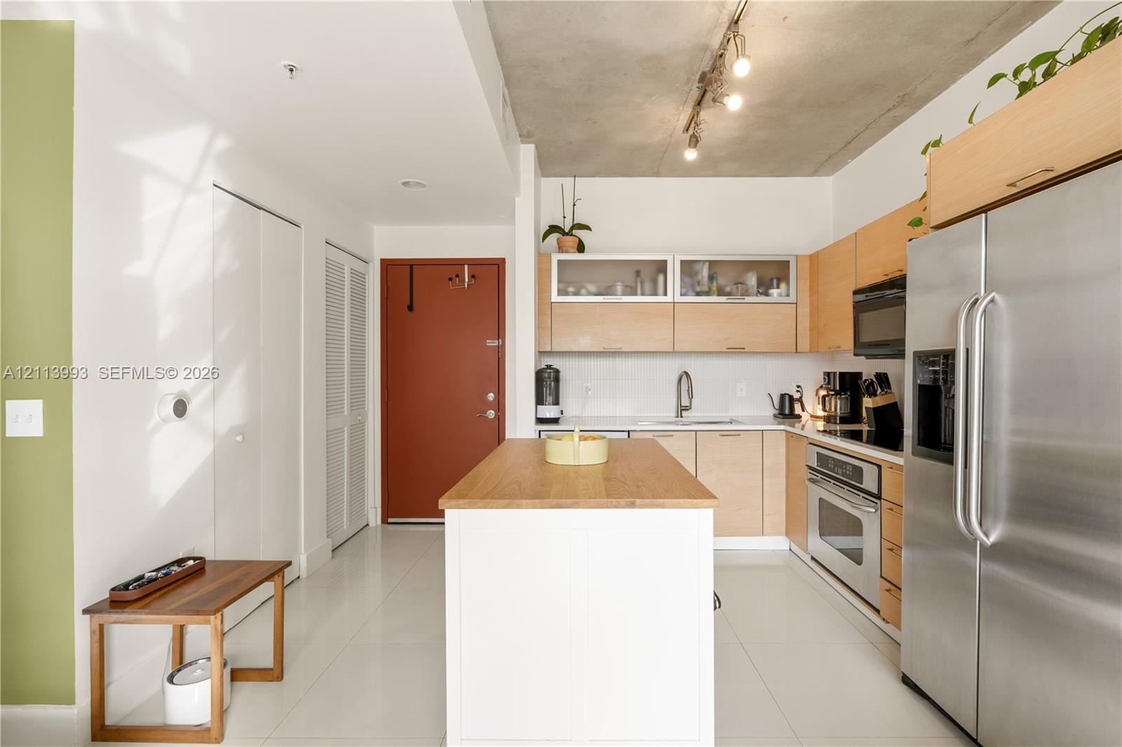 Kitchen featuring light-finish cabinetry with glass-front uppers, a central island with a wood countertop, exposed concrete ceilings with track lighting, stainless steel appliances, and tile flooring