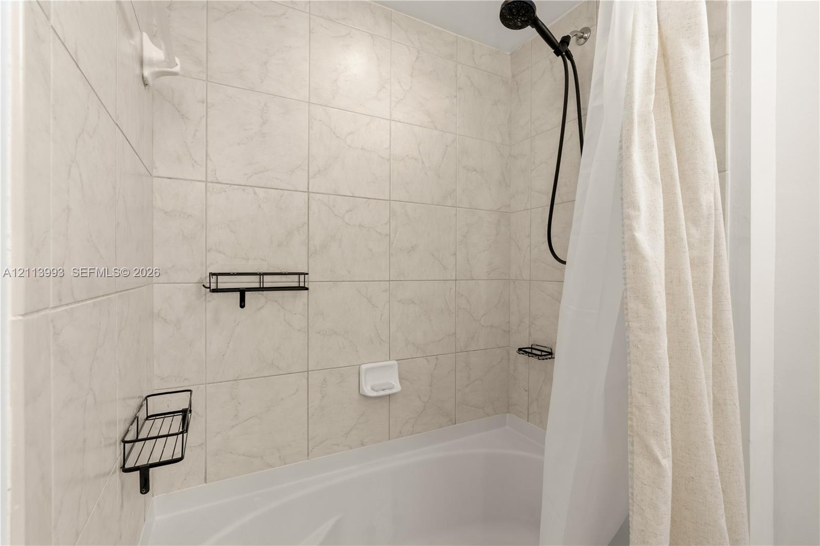 Bathtub enclosure featuring neutral tile wall surround, black finish handheld shower head, and dual black wire wall-mounted storage caddies