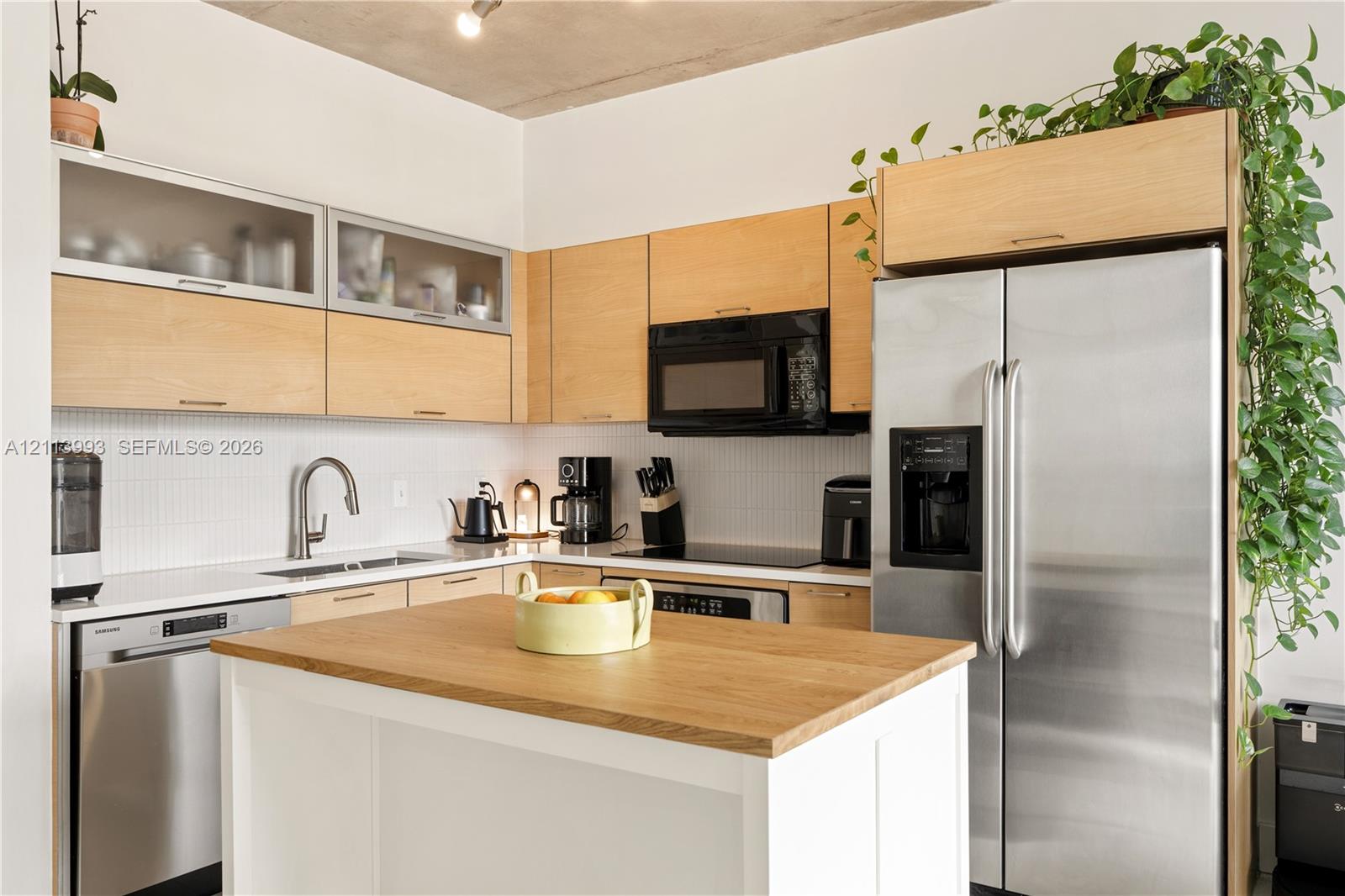 Kitchen featuring light wood-finish flat-panel cabinetry with glass-front uppers, white backsplash tile, stainless steel appliances, and a center island with a wood-finish countertop