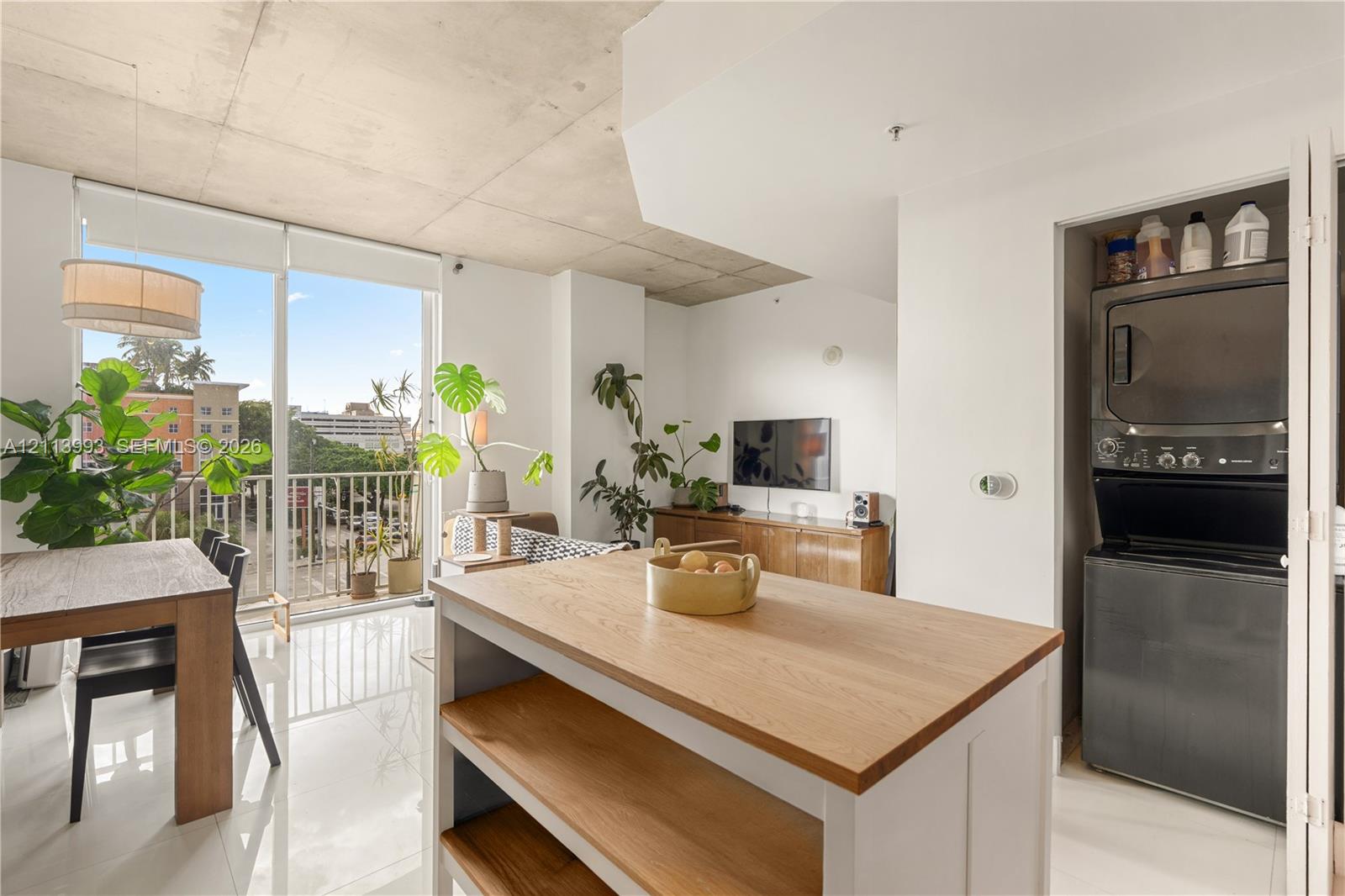 Open-concept interior featuring high exposed concrete ceilings, floor-to-ceiling glass paneling, polished tile flooring, a kitchen island with a wood countertop, and a built-in stackable laundry unit