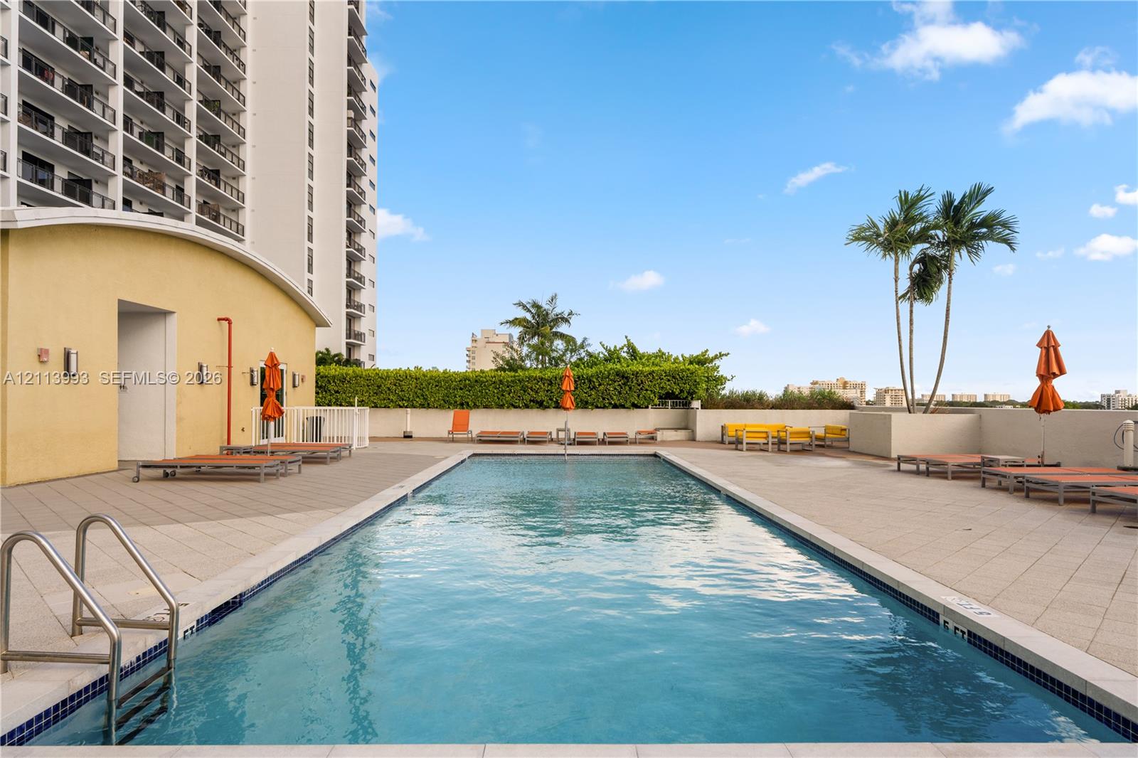 Swimming pool with tiled decking, metal handrails, and a surrounding perimeter wall with greenery