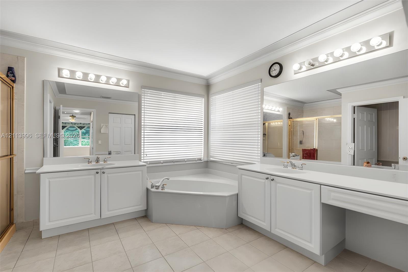 Dual white cabinetry vanities with inset-panel doors, solid surface countertops, corner soaking tub, ceramic tile flooring, and oversized wall-to-wall mirrors