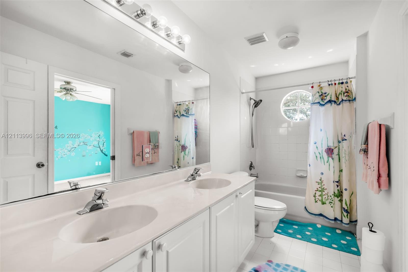 Double vanity with integrated white countertops and dual under-mount sinks, expansive wall-to-wall vanity mirror, overhead vanity lighting, circular accent window, and white tile tub surround