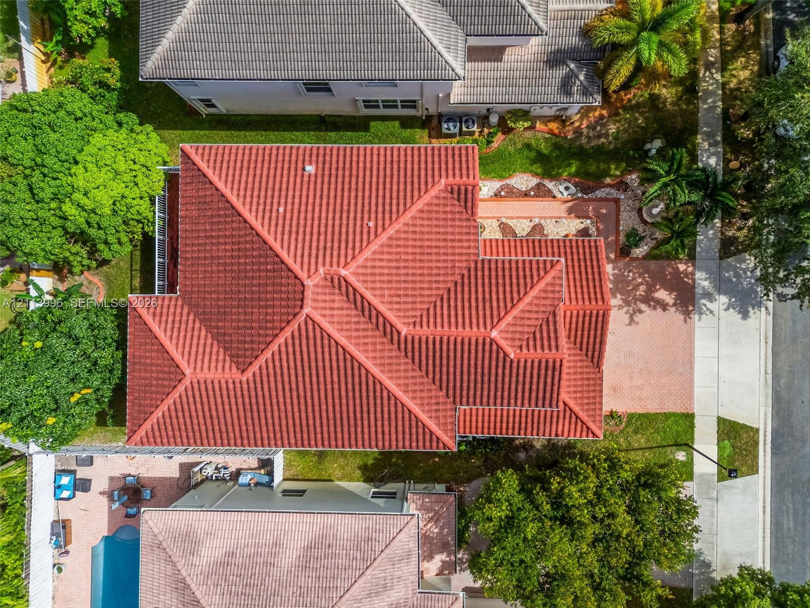 Aerial perspective of a multi-hip tile roof, expansive brick-paver driveway, and landscaped grounds featuring mature trees and established shrubbery