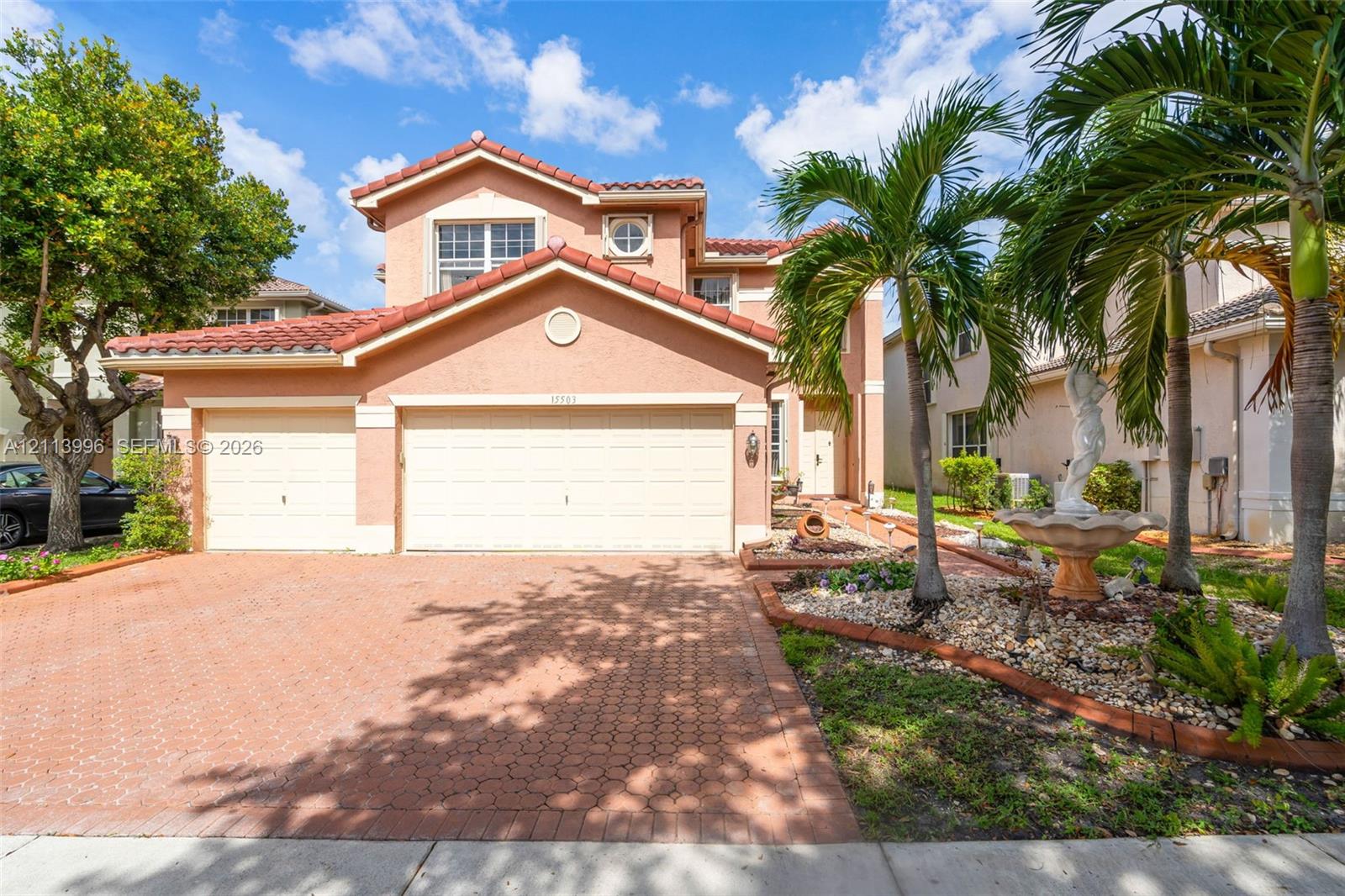 Two-story stucco exterior featuring a clay tile roof, two-car garage, paved driveway, and decorative stone-bordered landscaping with a pedestal fountain