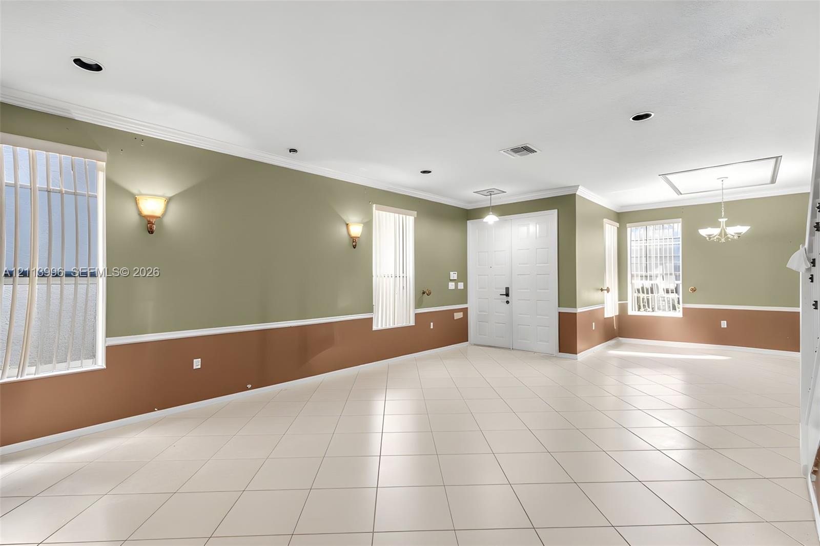 Open-concept interior featuring light tile flooring, crown molding, double-door entryway, dual wall-mounted sconce lighting, and a multi-arm chandelier