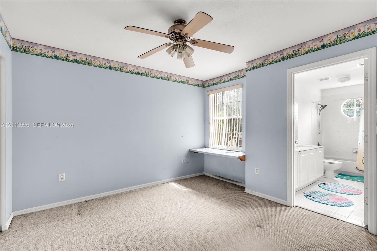 Room featuring neutral carpeting, a ceiling fan with light fixture, a built-in wall desk beneath a window, and a doorway to a bathroom