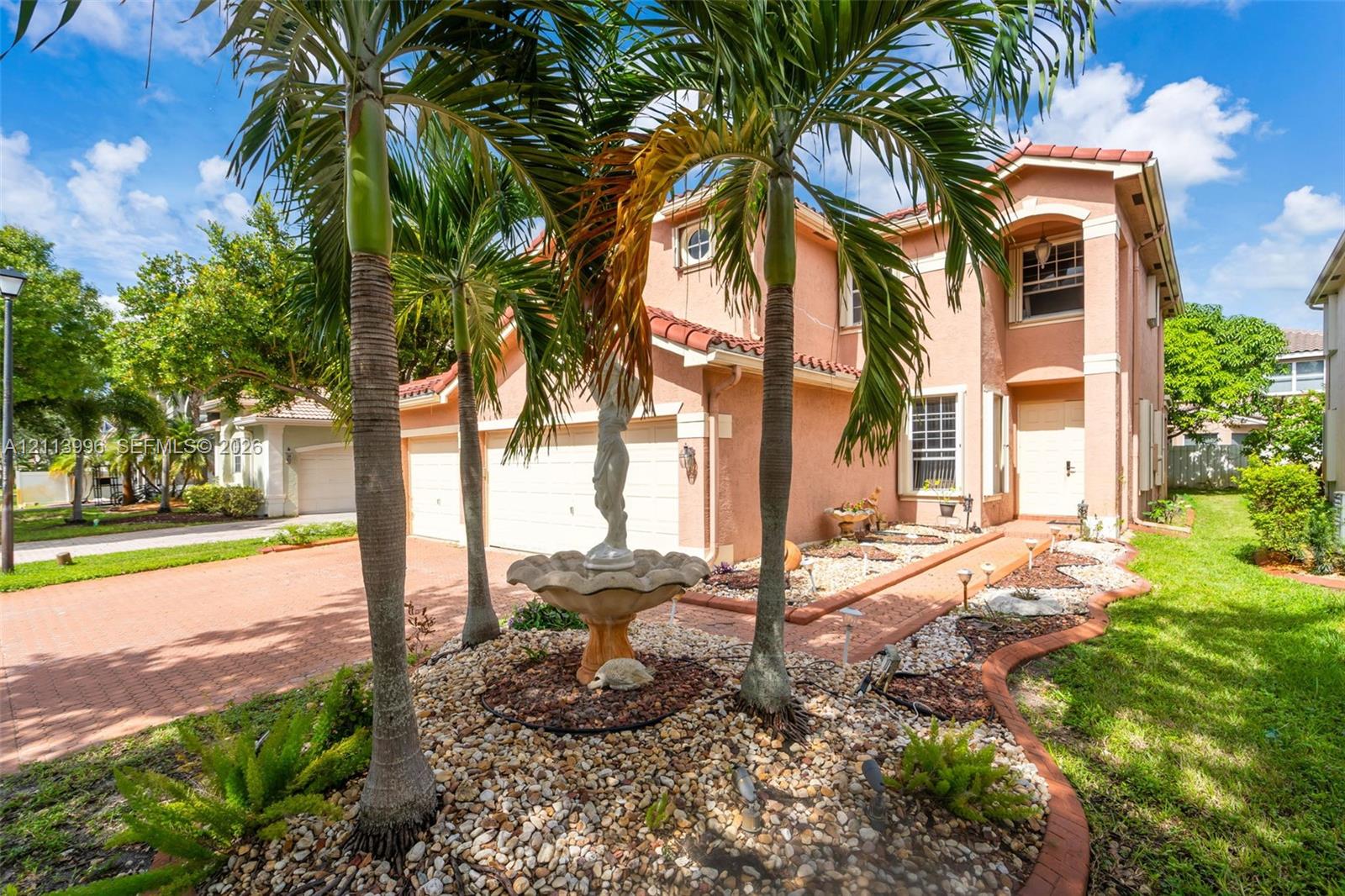 Two-story exterior featuring a stucco facade, red tile roof, multi-car garage, paver driveway, and front garden with a central fountain