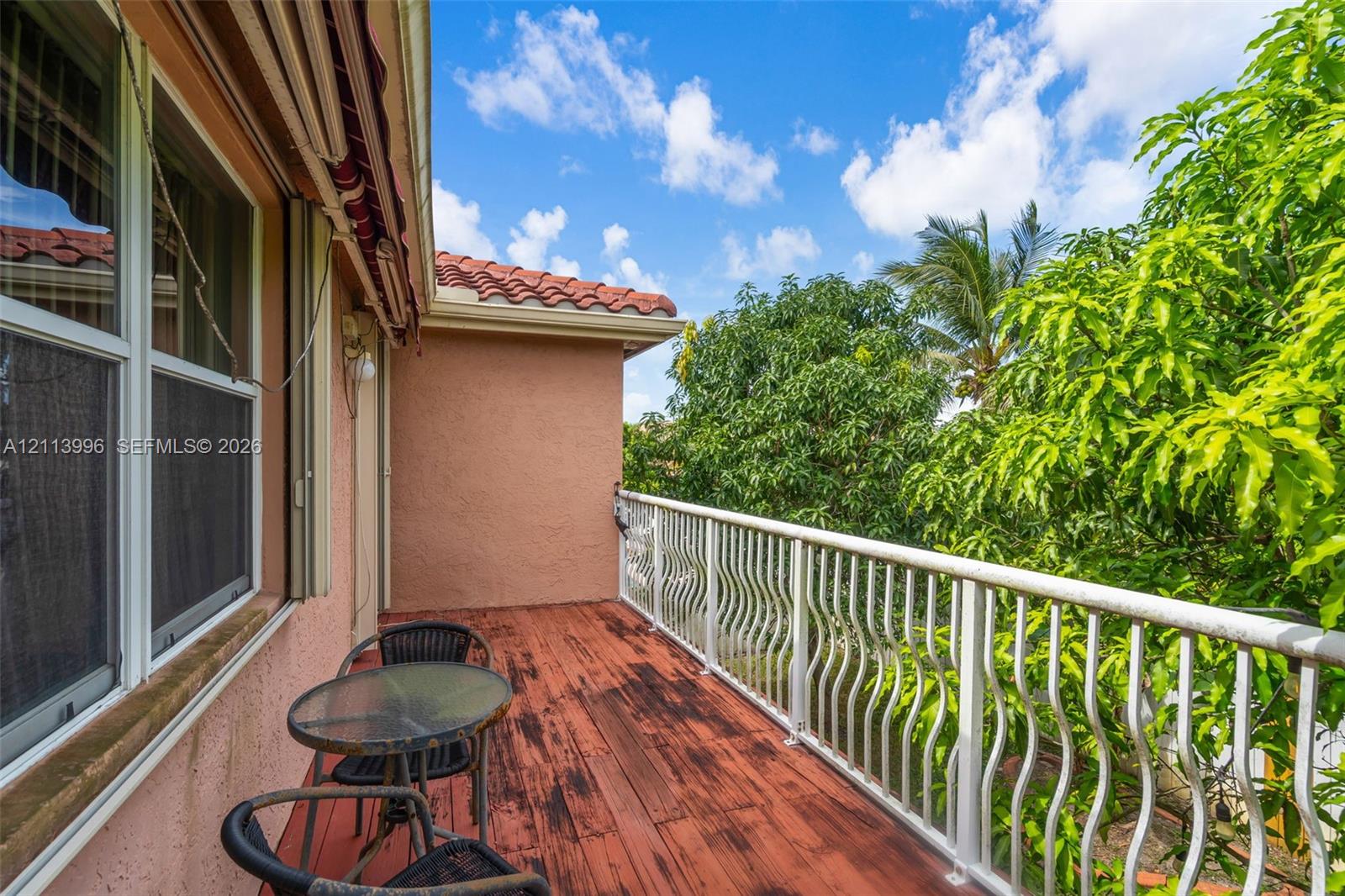 Exterior balcony featuring red-toned wood-finish decking, white metal balustrades, stucco wall cladding, and a red tile roofline