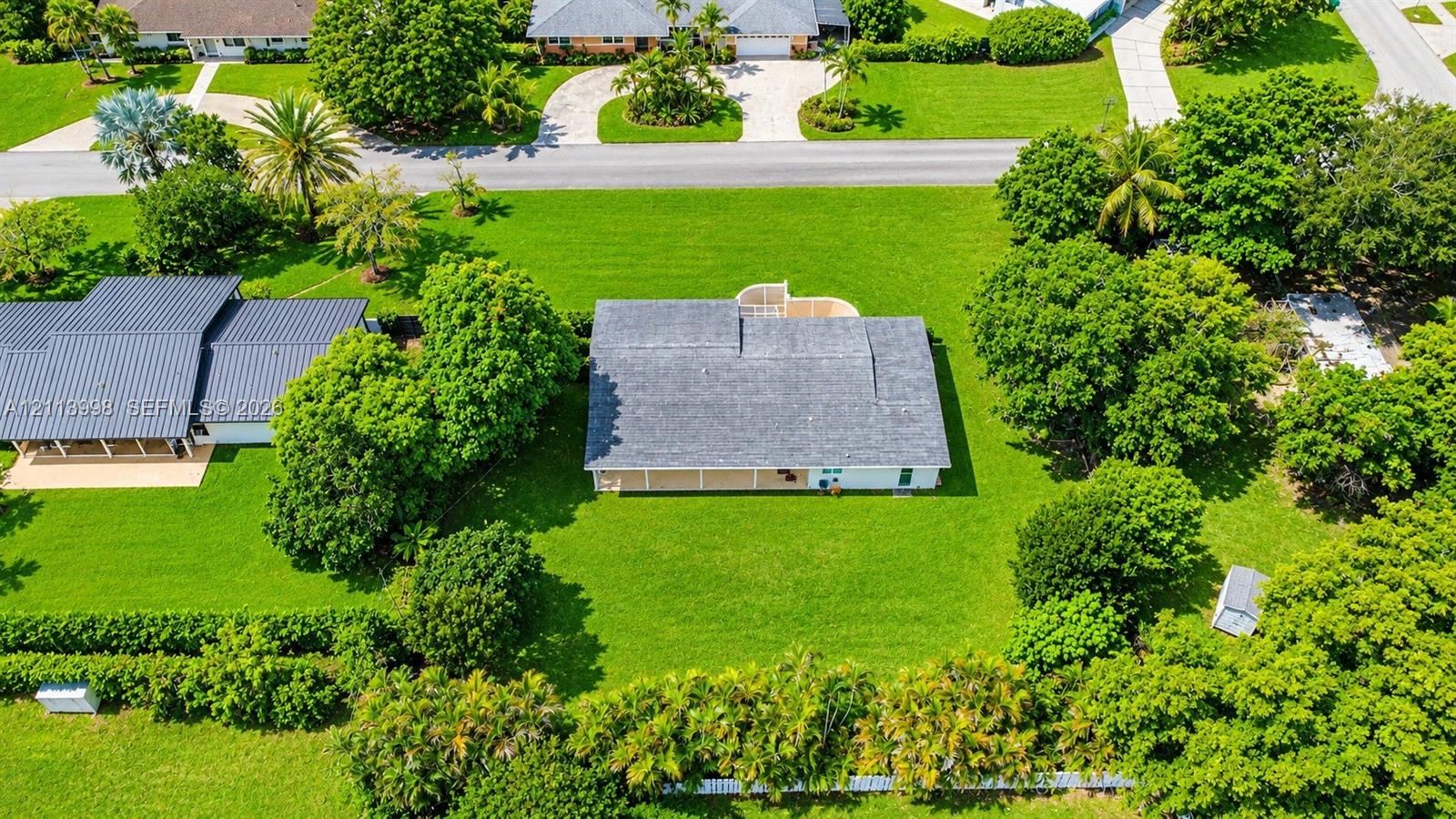 Aerial perspective featuring a single-story residence with a shingle roof, integrated rear covered patio, expansive manicured lawn, and mature perimeter landscaping