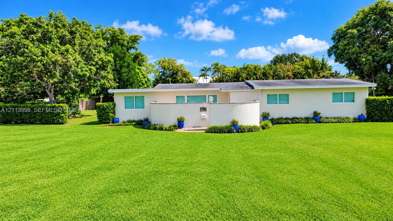 Single-story exterior featuring a stucco faade, a semi-circular front privacy wall, expansive manicured lawn, and mature perimeter landscaping