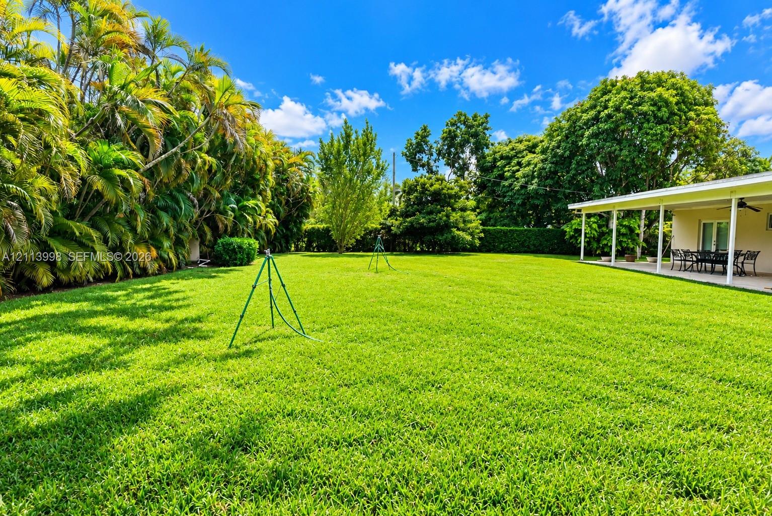 Expansive backyard featuring a manicured lawn, perimeter landscaping, mature trees, and a covered patio