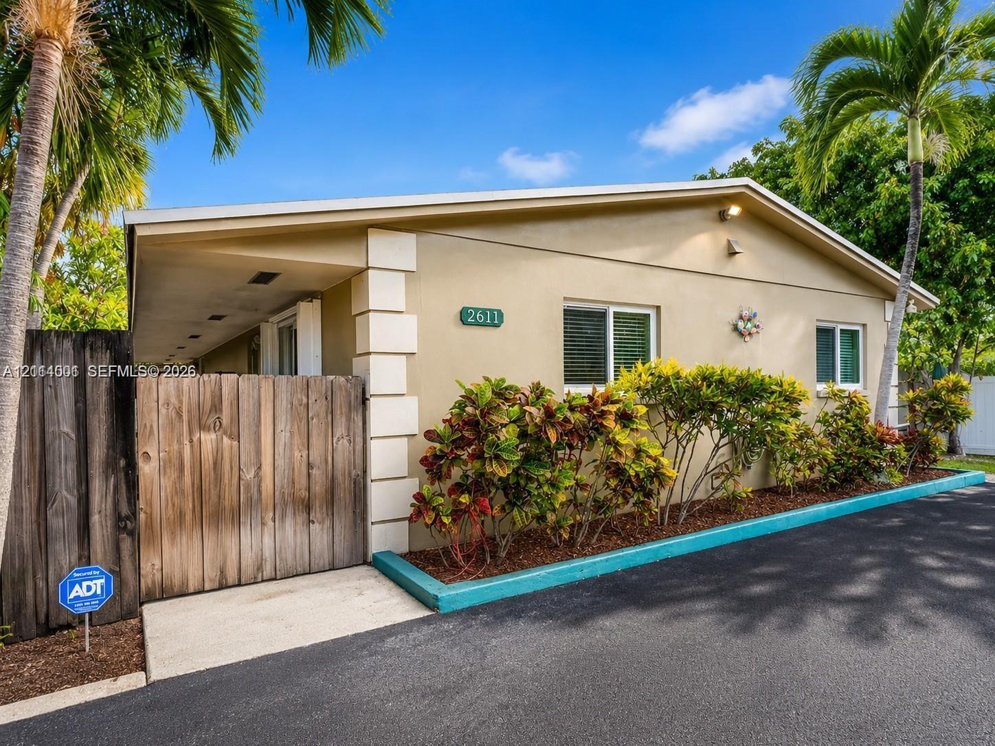 Stucco exterior featuring decorative corner quoins, a wood-slat privacy gate, asphalt driveway, and integrated landscaping with mature palm trees