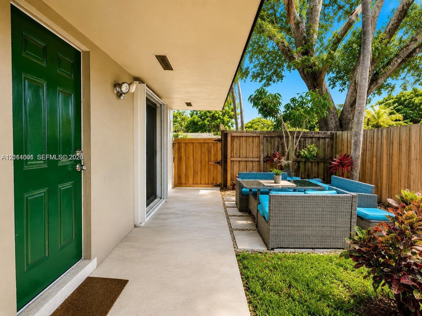 Covered concrete patio featuring a solid-color panel door, exterior wall-mounted lighting, and a wood privacy fence