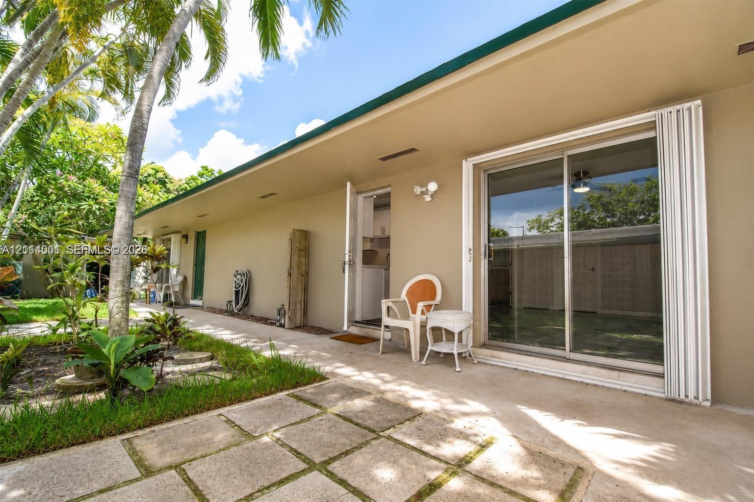 Concrete patio with square pavers, exterior sliding glass door, hinged entry door, and perimeter landscaping with palm trees