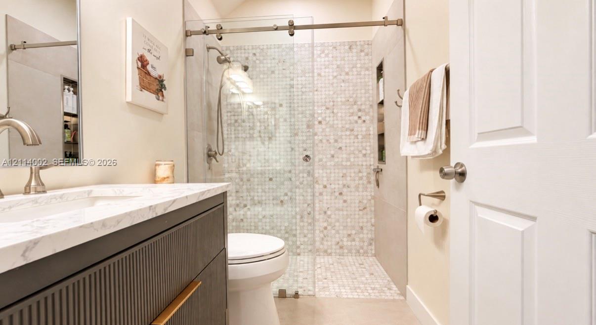 Bathroom featuring a fluted wood-finish vanity with a stone-surface countertop, an integrated sink, a glass-enclosed walk-in shower with mosaic tile accents, and built-in wall shelving