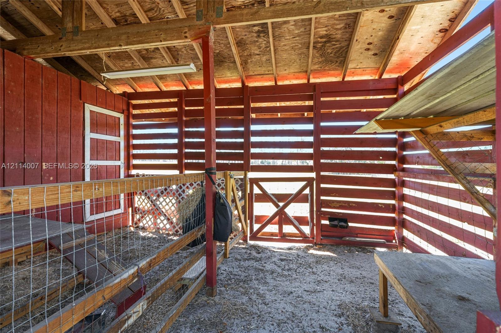 Open-air structure featuring red-painted horizontal wood siding, exposed rafter ceilings, wood-frame partitions, wire fencing, and an integrated wood-frame gate