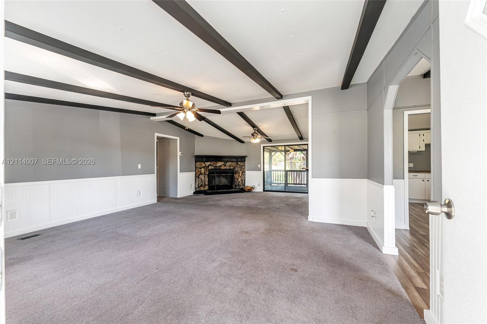 Living area featuring decorative ceiling beams, stone-surround fireplace, dual ceiling fans, and wainscoting