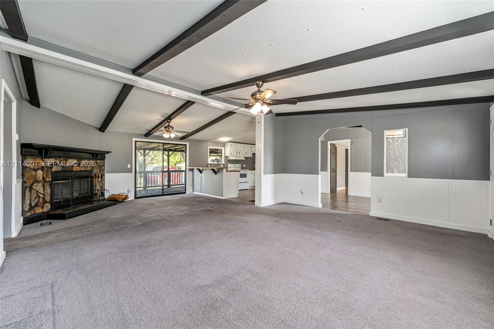 Vaulted ceiling featuring exposed dark beams, floor-to-ceiling stone fireplace, beadboard wainscoting, sliding glass door, and dual ceiling fans