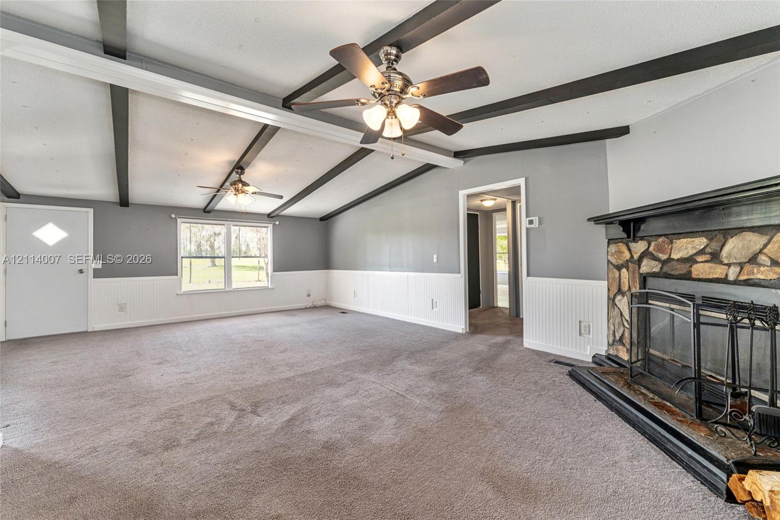 Vaulted ceiling with exposed decorative beams, stone-faced fireplace with a mantel, white wainscoting, and dual ceiling fans