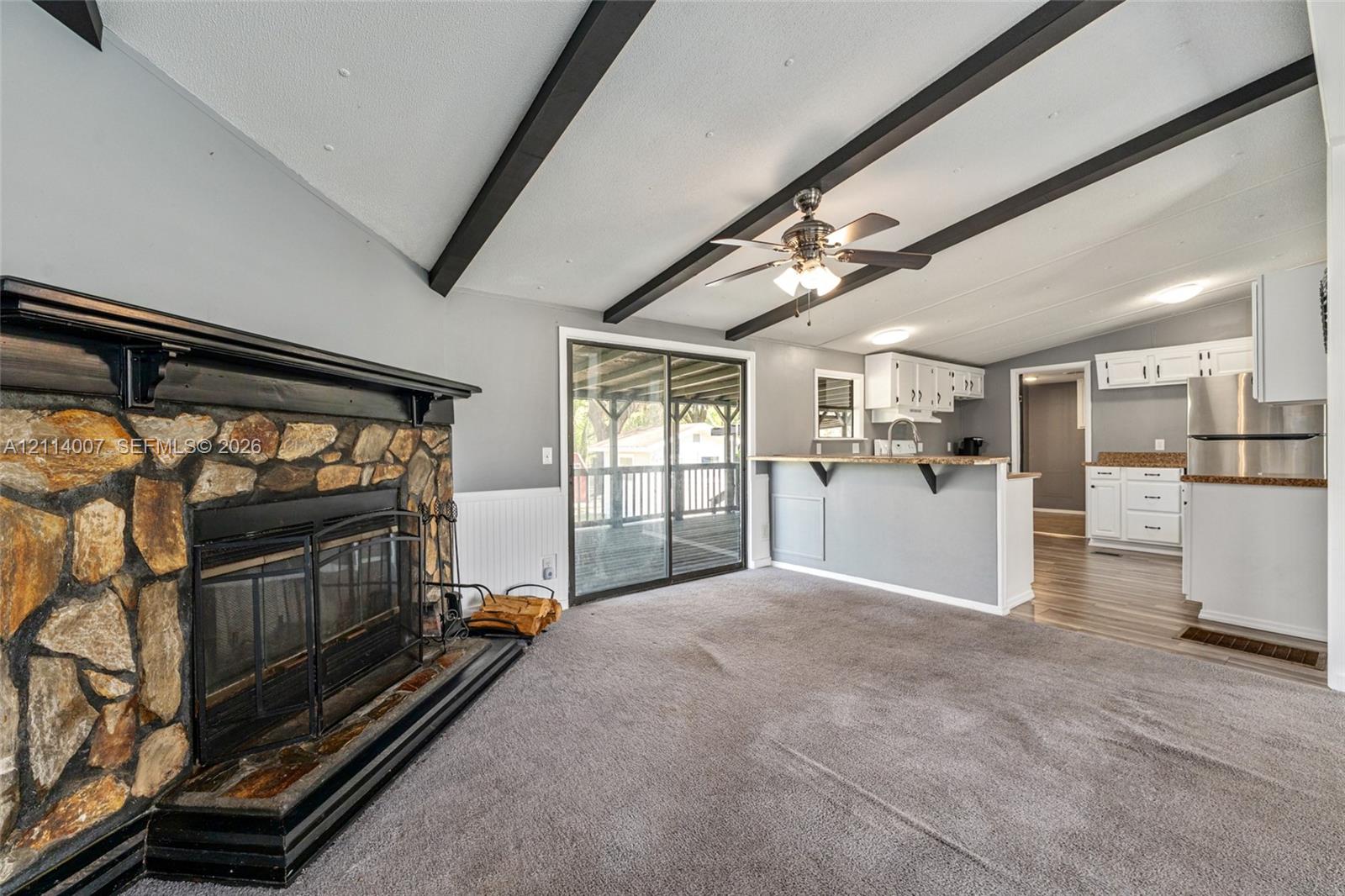 Vaulted ceiling featuring exposed dark beams, stone-faced fireplace with a black mantel, kitchen peninsula with overhead cabinetry, and sliding glass doors