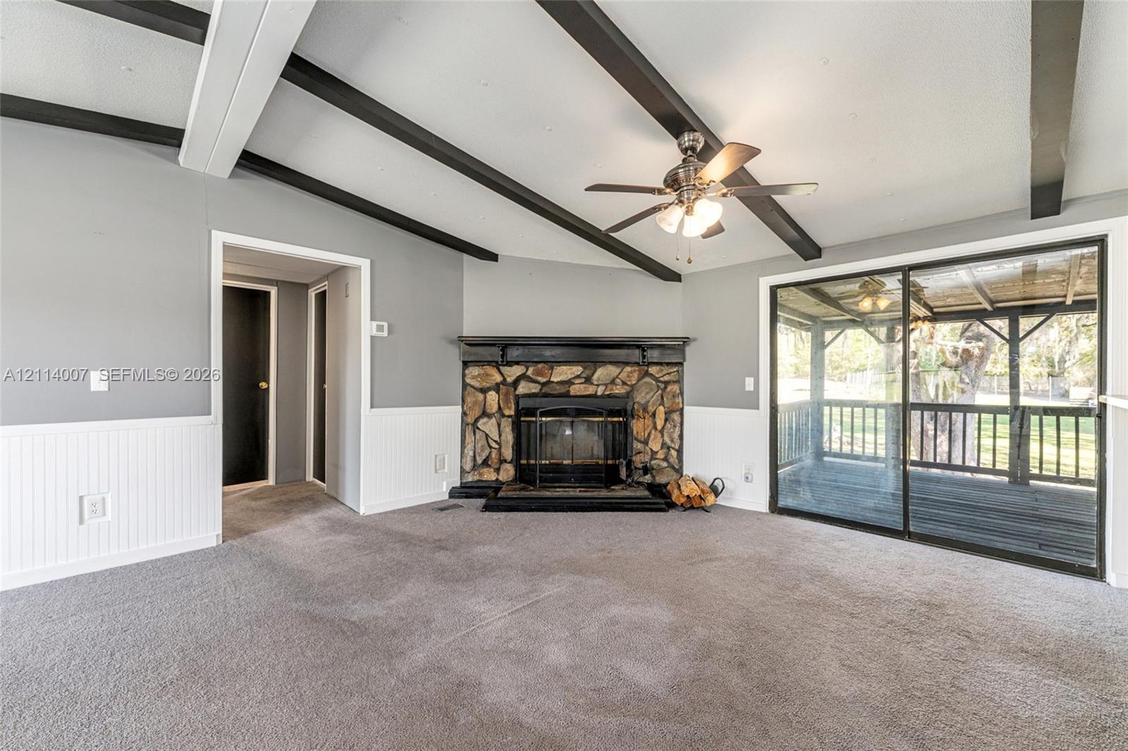 Living area featuring vaulted ceilings with decorative exposed beams, stone-surround fireplace with a wooden mantel, white wainscoting, and sliding glass doors