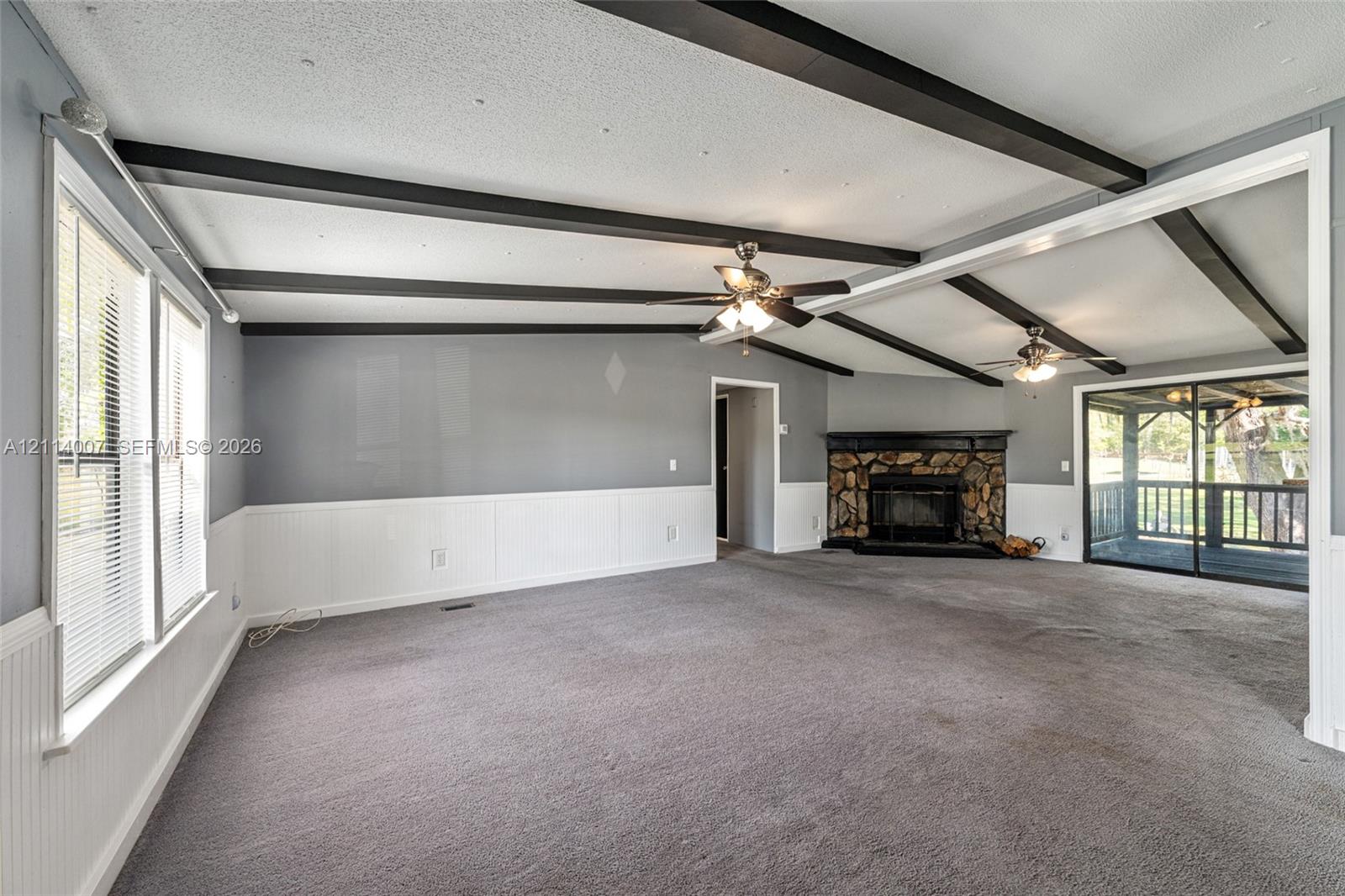 Living area featuring exposed ceiling beams, a stone-surround fireplace, wainscoting, and dual ceiling fans