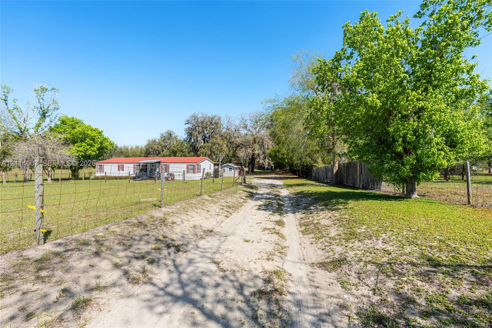 Single-level dwelling with a red metal roof, gravel driveway, wire fencing, and mature trees on a sprawling lot