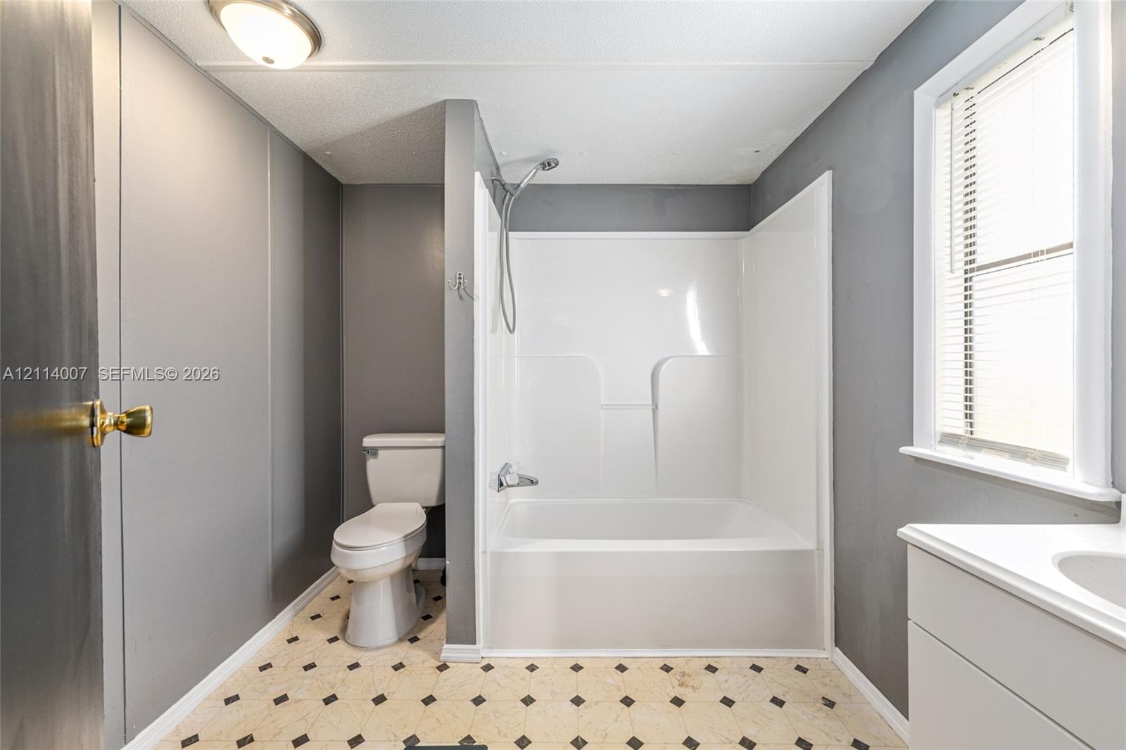 Bathroom featuring a white tub-shower combination, pedestal toilet, white vanity with an integrated sink, window with horizontal blinds, and patterned tile flooring
