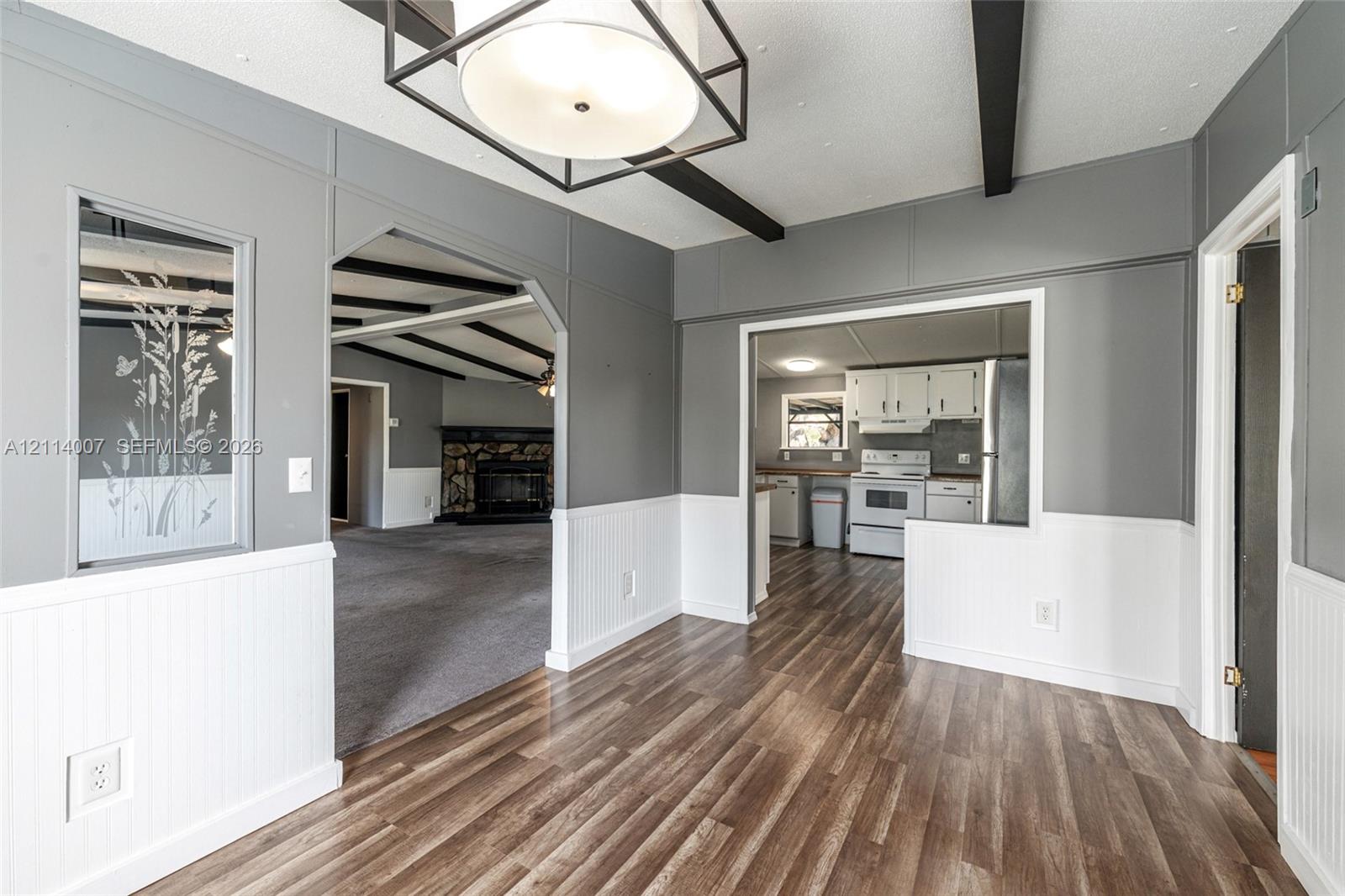 Dining area featuring wood-finish flooring, beadboard wainscoting, a geometric lantern-style pendant light fixture, and dark exposed ceiling beams