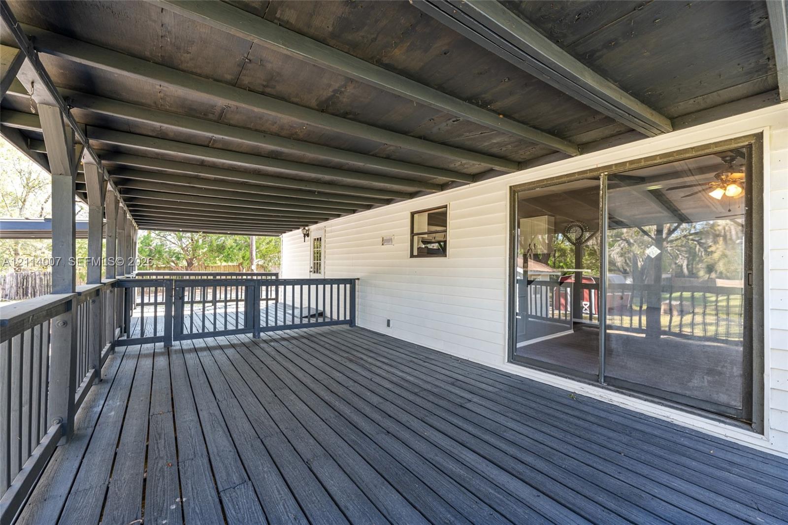 Covered wooden deck featuring dark-stained plank flooring, an overhead beam structure, perimeter railing with a gated entrance, and a sliding glass door