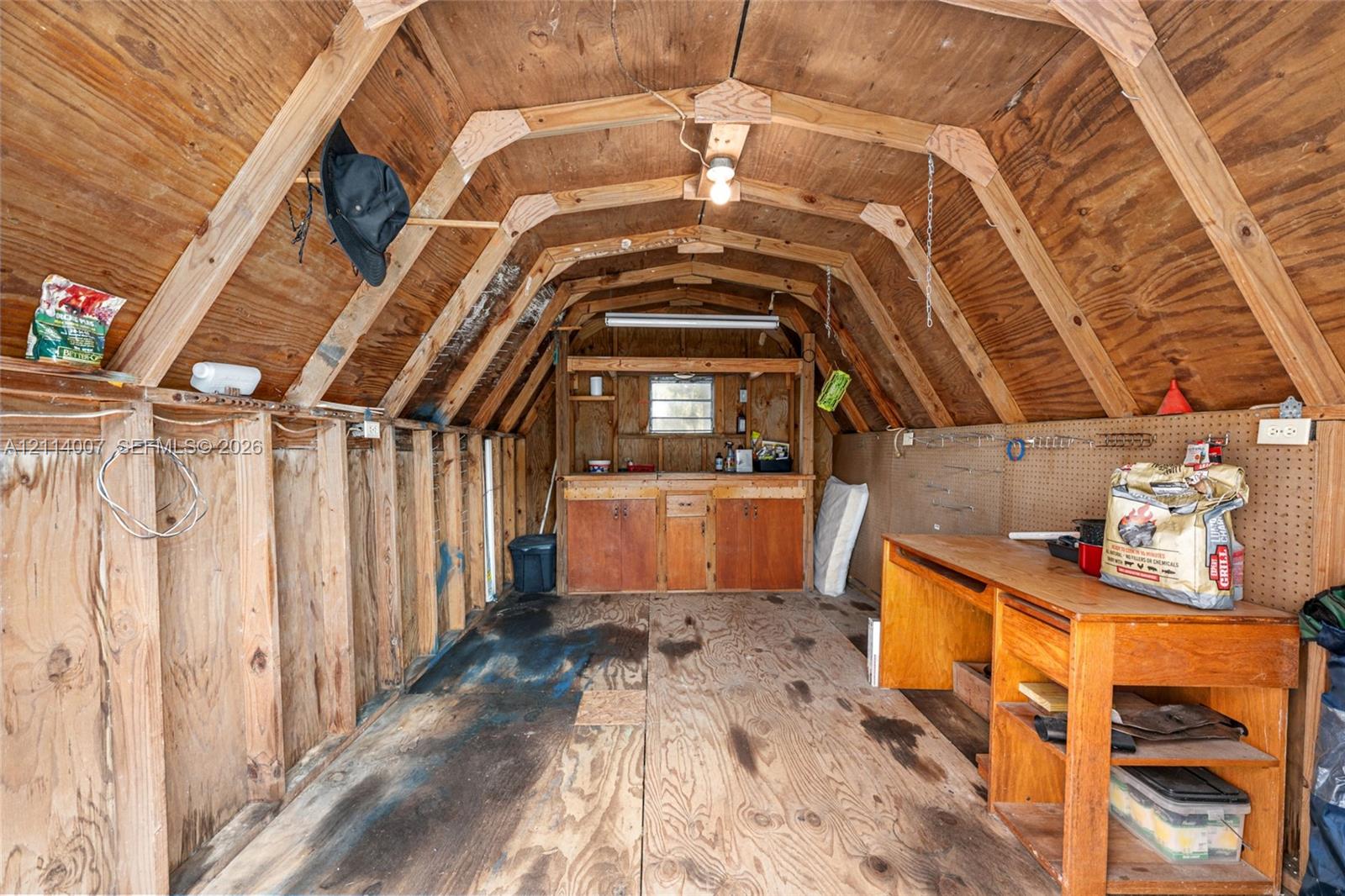 Detached storage structure featuring exposed wood framing, plywood wall paneling, built-in wooden cabinetry, integrated workbench, and overhead lighting